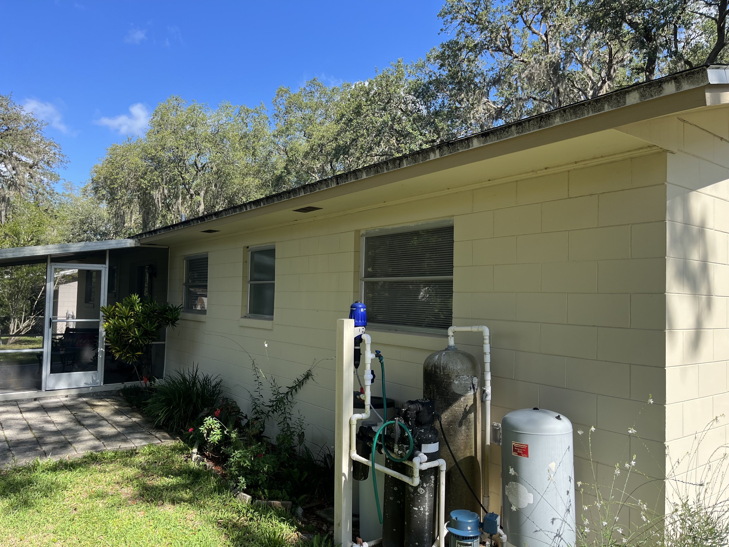 A side of a yellow house with windows, surrounded by greenery and trees, with a water filtration or pump system outside.