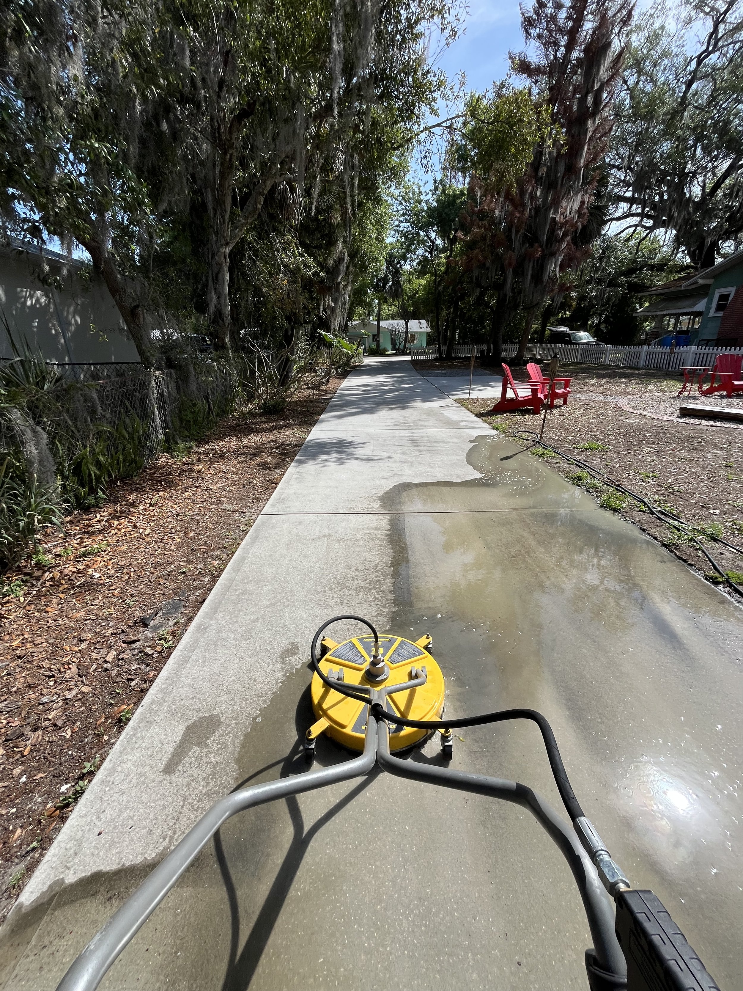 View from a sidewalk or driveway of a yellow pressure washer with a circular yellow spray head on the front, positioned on the concrete path. The path is wet with some puddles, and the surroundings include trees, bushes, and neighboring houses with w