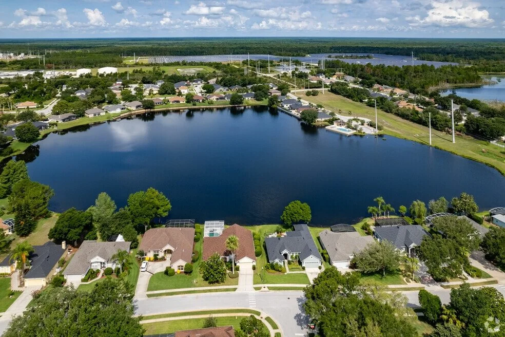 Aerial view of a residential neighborhood surrounding a large pond with houses, trees, and greenery, with a water body and open fields in the distance under a partly cloudy sky.