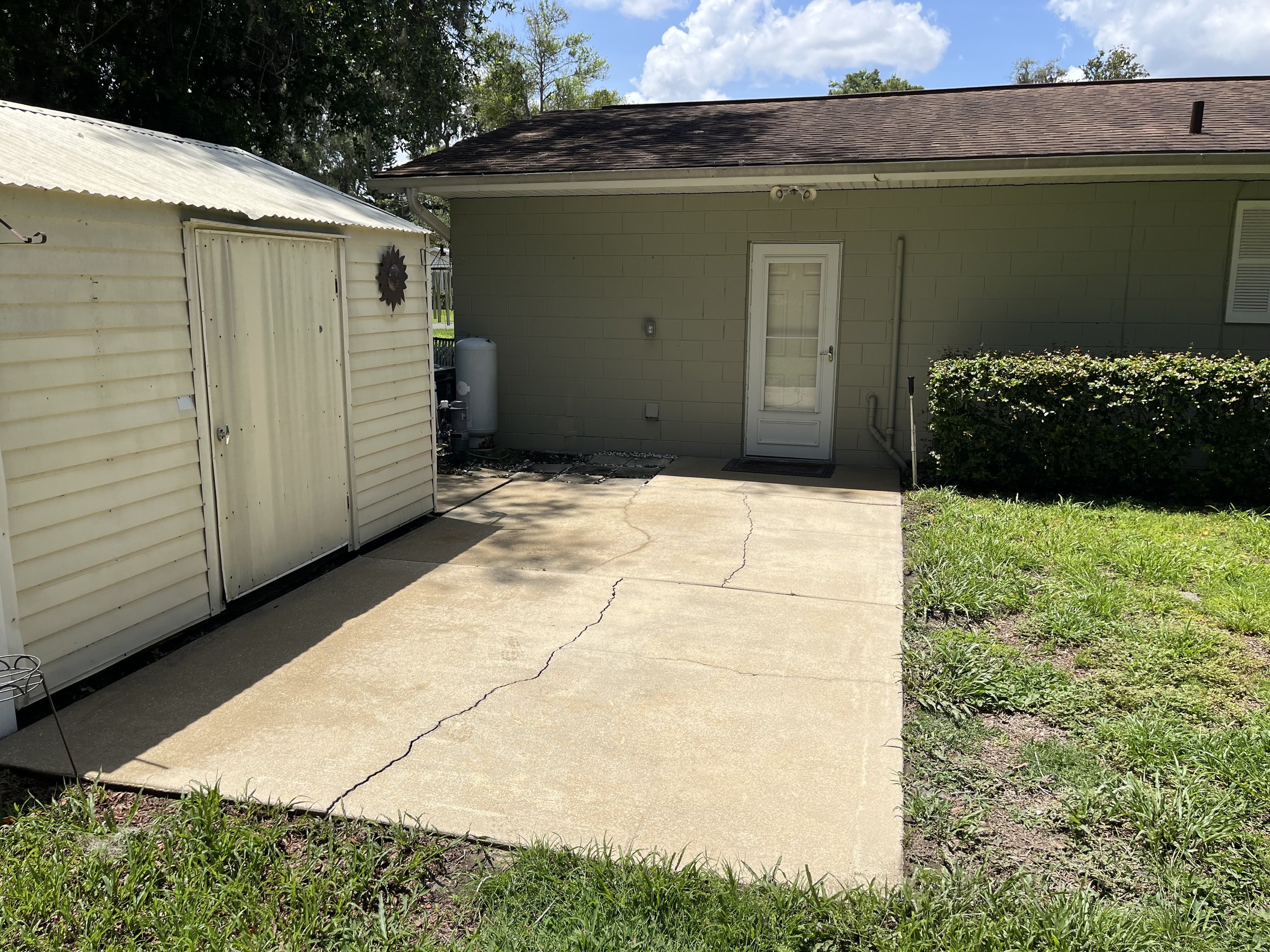 A backyard area with a concrete patio, a white shed on the left, a light green house with a door in the middle, and a small bush on the right. There are trees and a partly cloudy sky in the background.