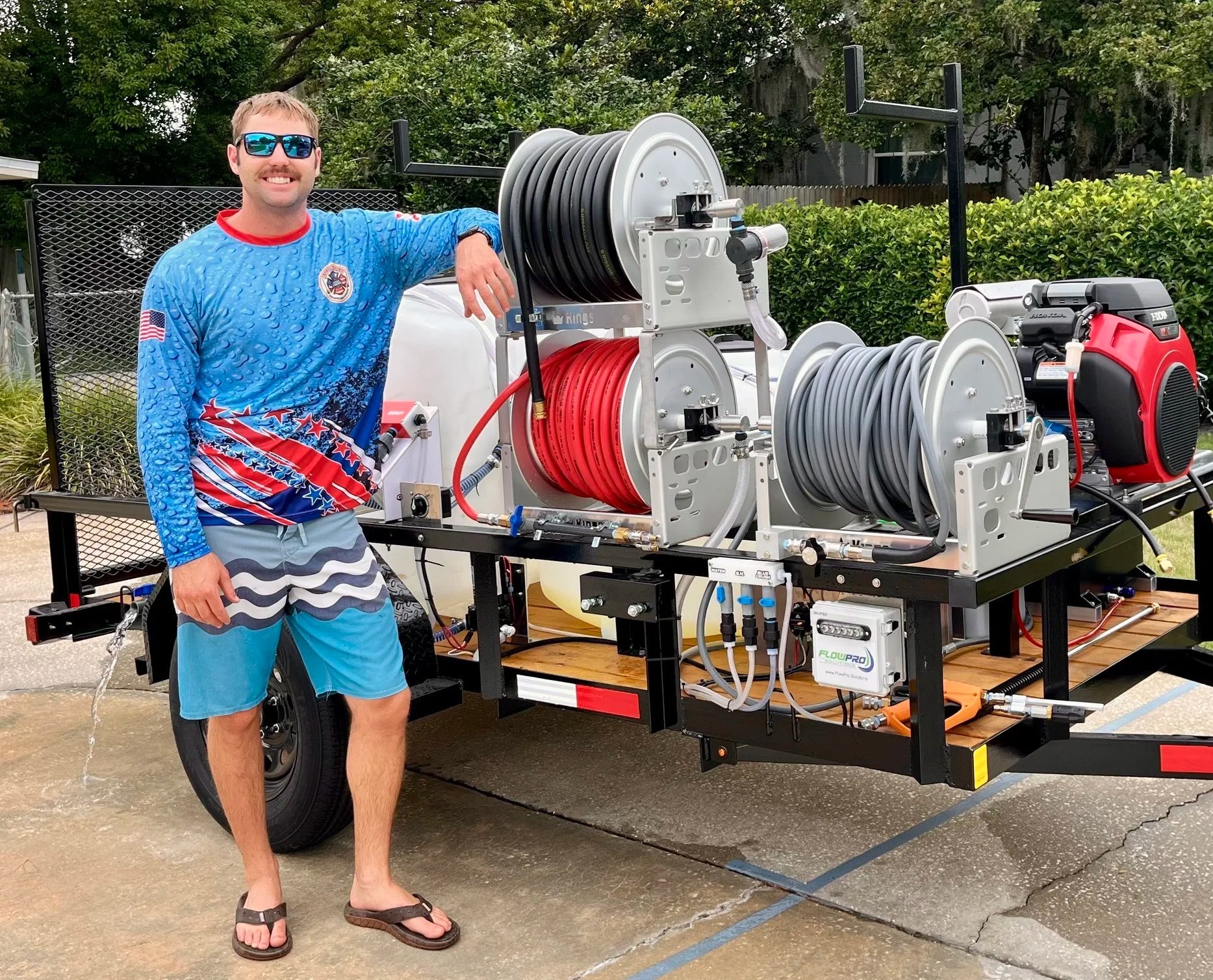 A man in swim trunks and a blue patriotic shirt standing next to a large portable fire suppression system on a trailer. He's wearing sunglasses and smiling.