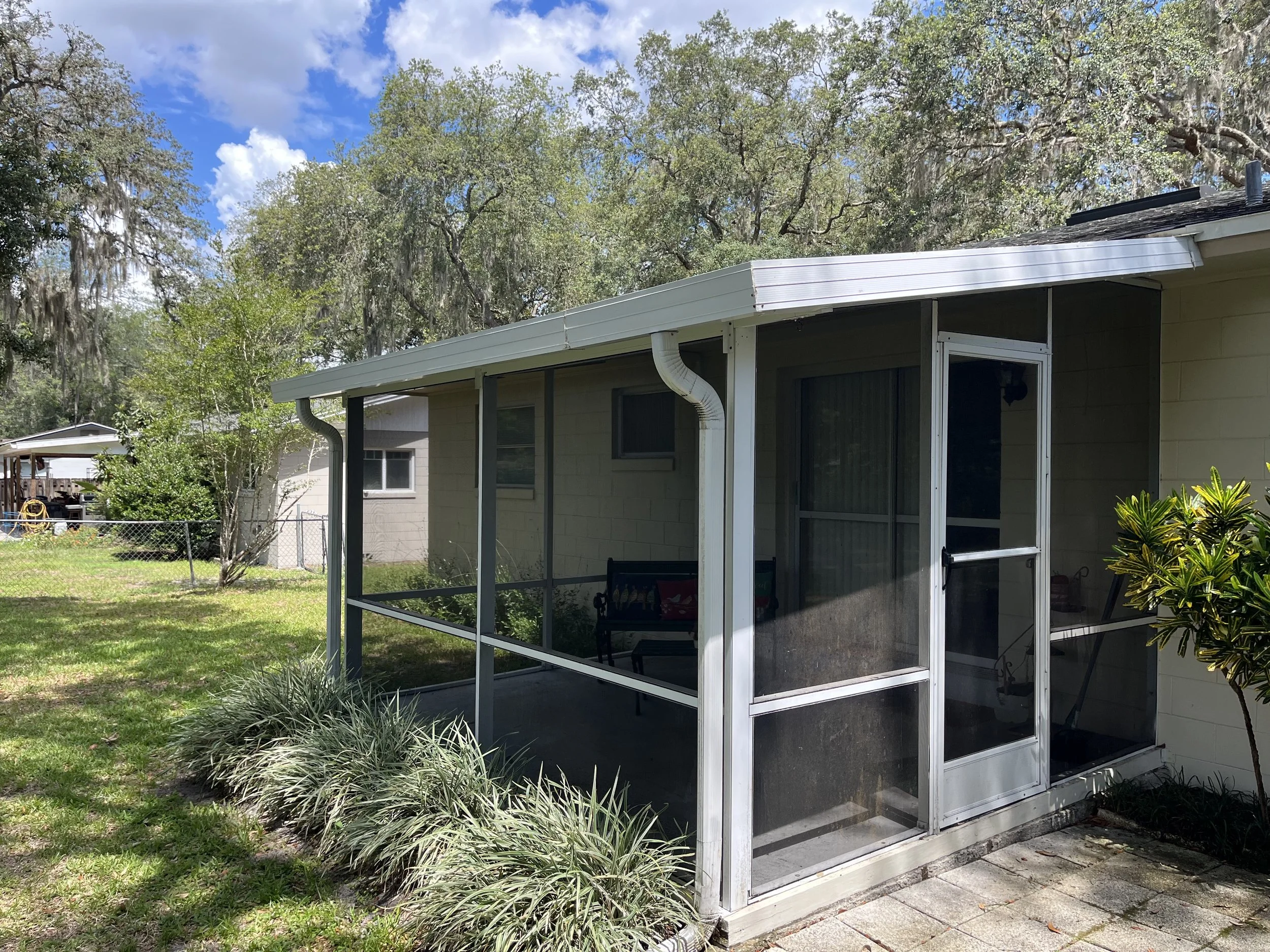 Backyard view with screened porch, trees, grass, and blue sky with clouds.