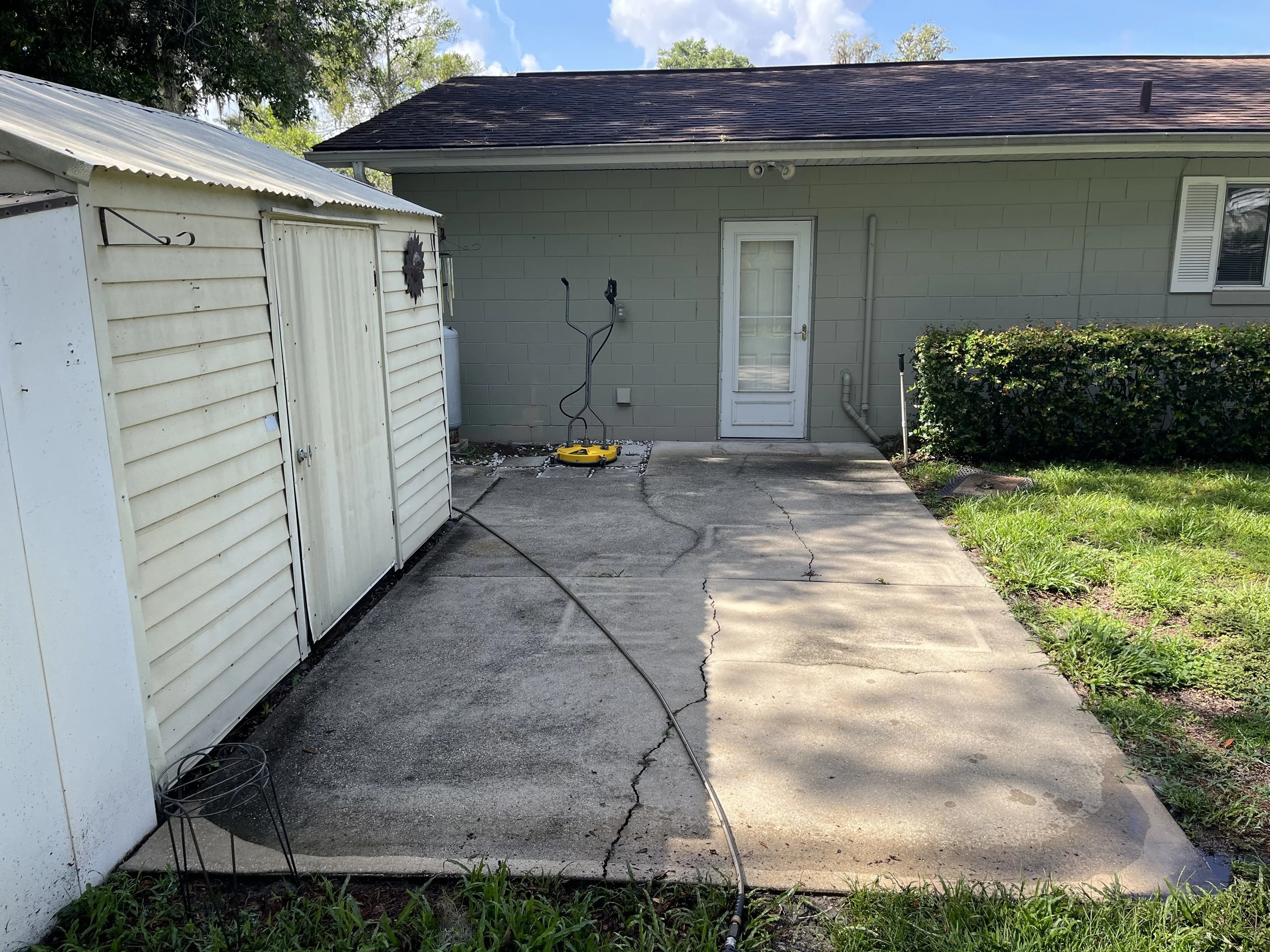 Backyard patio with concrete surface, a white shed on the left, a light green house with window and door in the center, some greenery including bushes and grass, and a blue sky with clouds overhead.