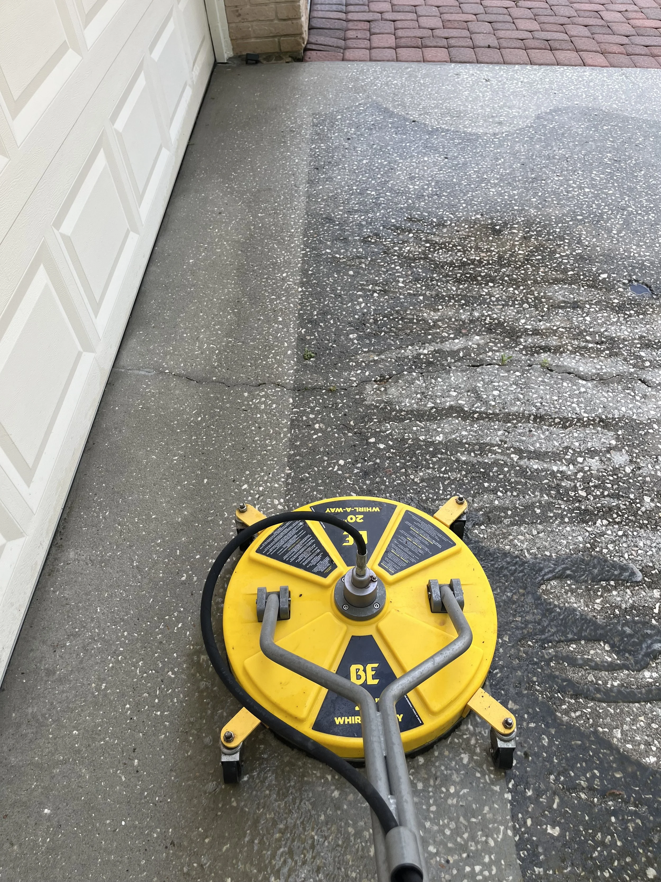 Overhead view of a yellow whirly tumbler cleaner on a concrete driveway in front of a white garage door and brick pavement.