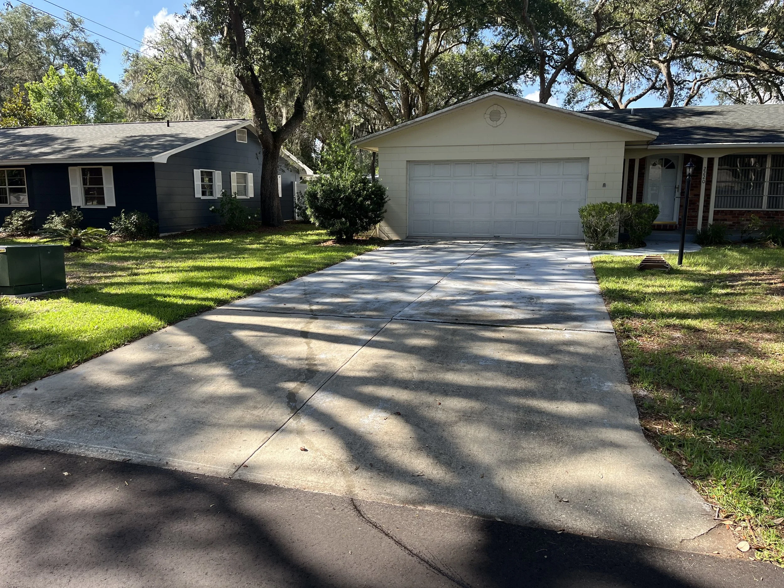 A residential neighborhood with a concrete driveway leading to a white garage door, flanked by green grass and bushes, with a house to the right and trees overhead casting shadows.