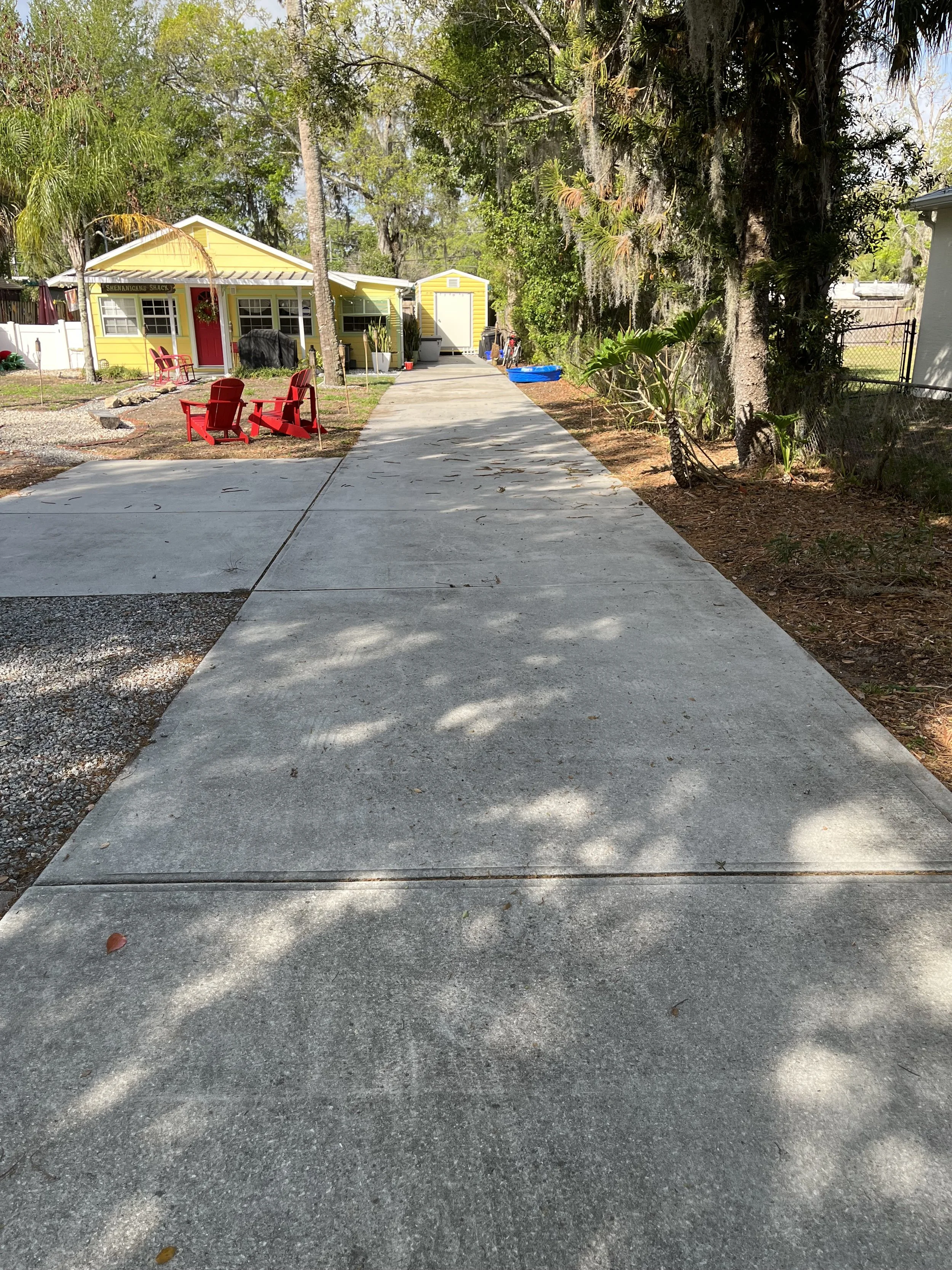 A concrete driveway leads to a yellow house with red door and red chairs outside. Tall trees and lush greenery surround the house, with a small shed nearby and a blue kiddie pool on the side.