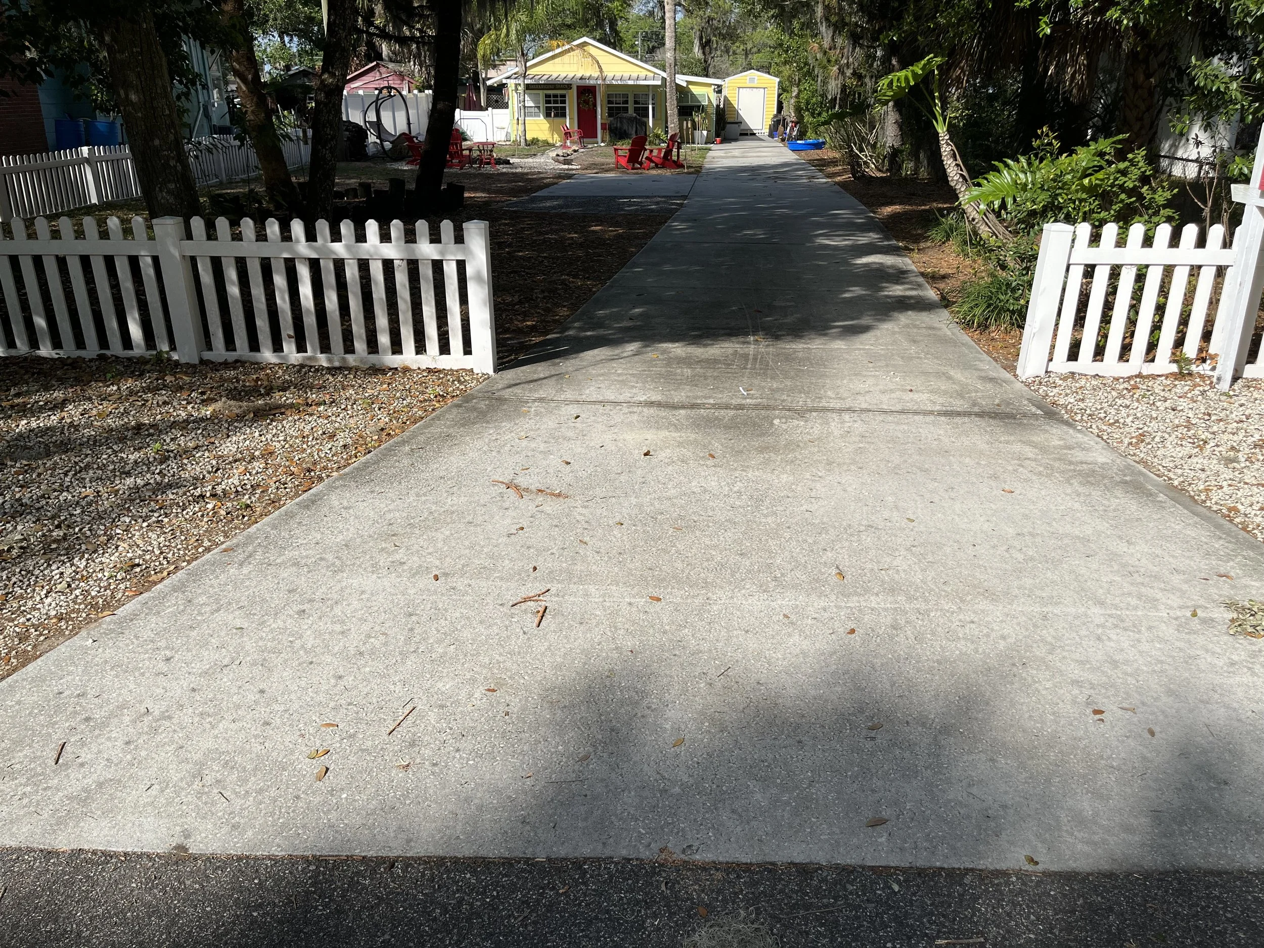 A sidewalk leading to a yellow house with a porch and red chairs, surrounded by trees and greenery, with a white picket fence on either side.