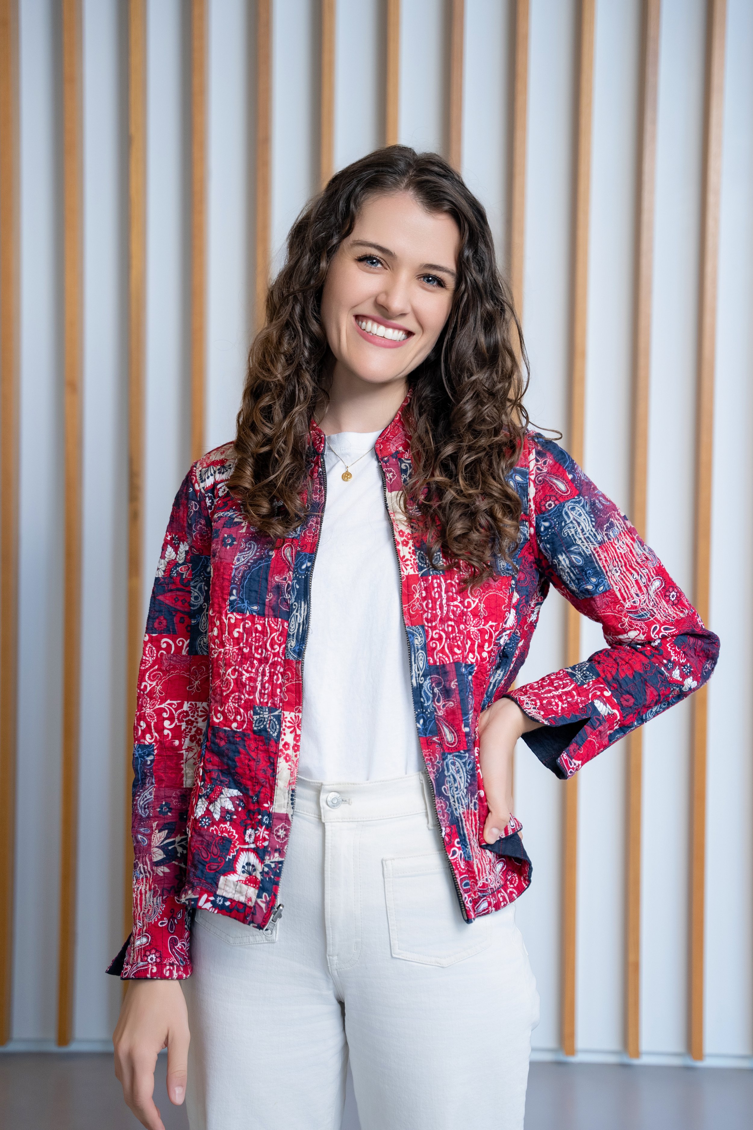 A young woman with long curly hair and blue eyes smiling, wearing a colorful patchwork jacket over a white shirt and white pants, standing in front of a wooden slat wall.