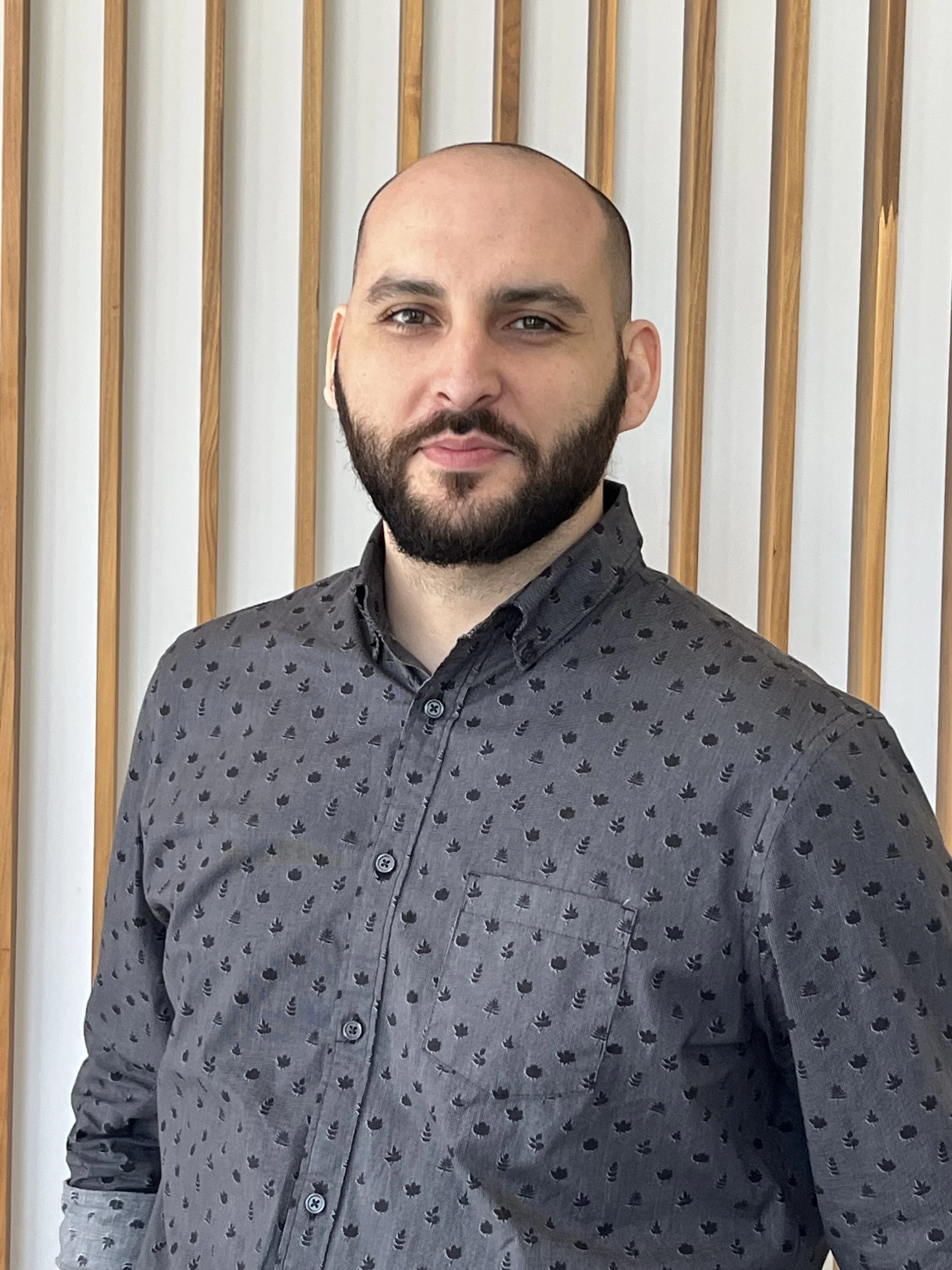 A man with a shaved head, beard, and mustache wearing a dark patterned button-up shirt, standing in front of a background with vertical wooden slats.