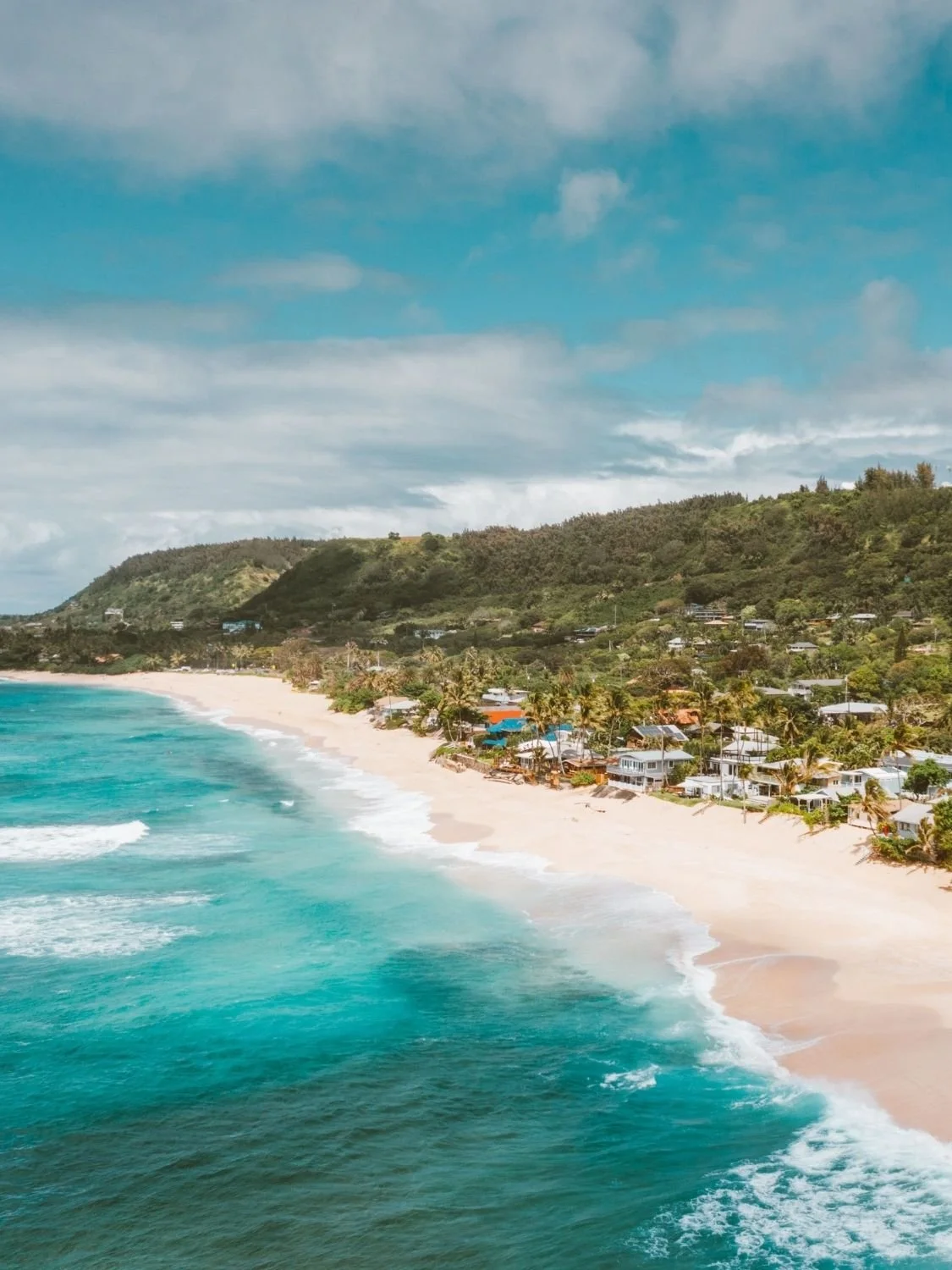 Aerial view of a tropical beach with turquoise ocean waves, golden sand, and beachfront homes backed by lush green hills.