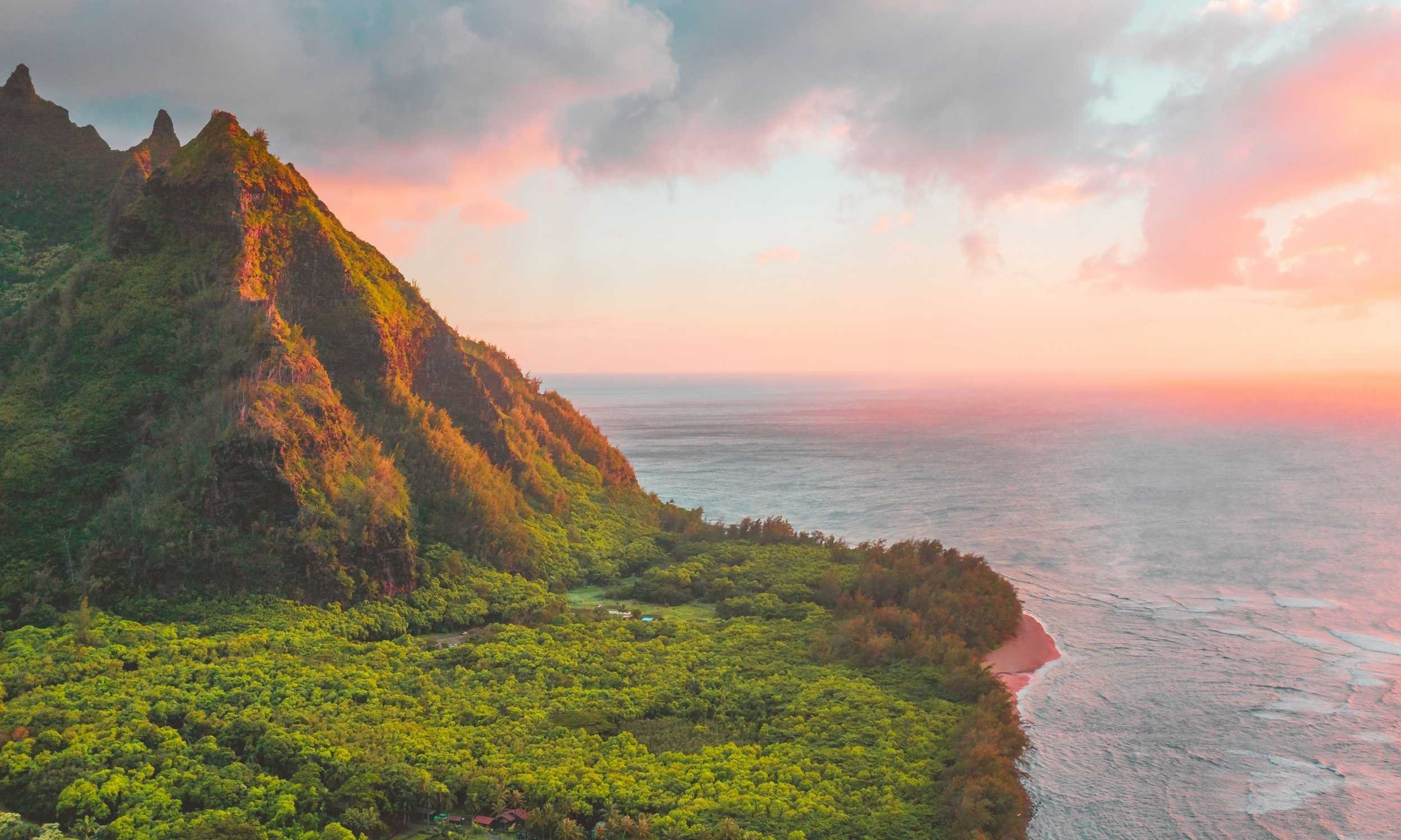 A dramatic green mountain rises above a forested coastline overlooking the ocean at sunset with colorful clouds overhead.