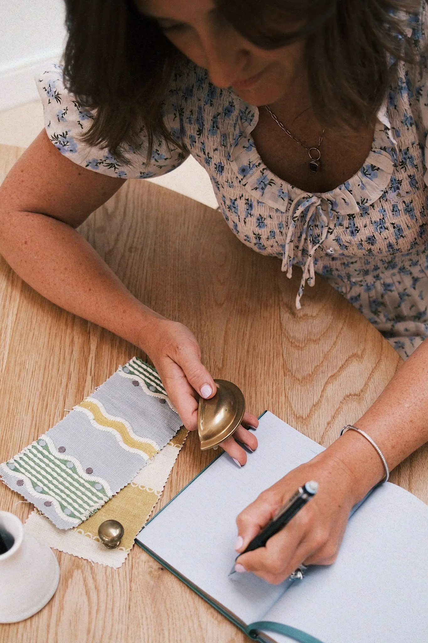 A woman is using a brass stamp to press ink onto fabric swatches, which are laid out on a wooden table. She is wearing a floral blouse and has accessorized with a necklace and a bracelet, and she is taking notes in a notebook.