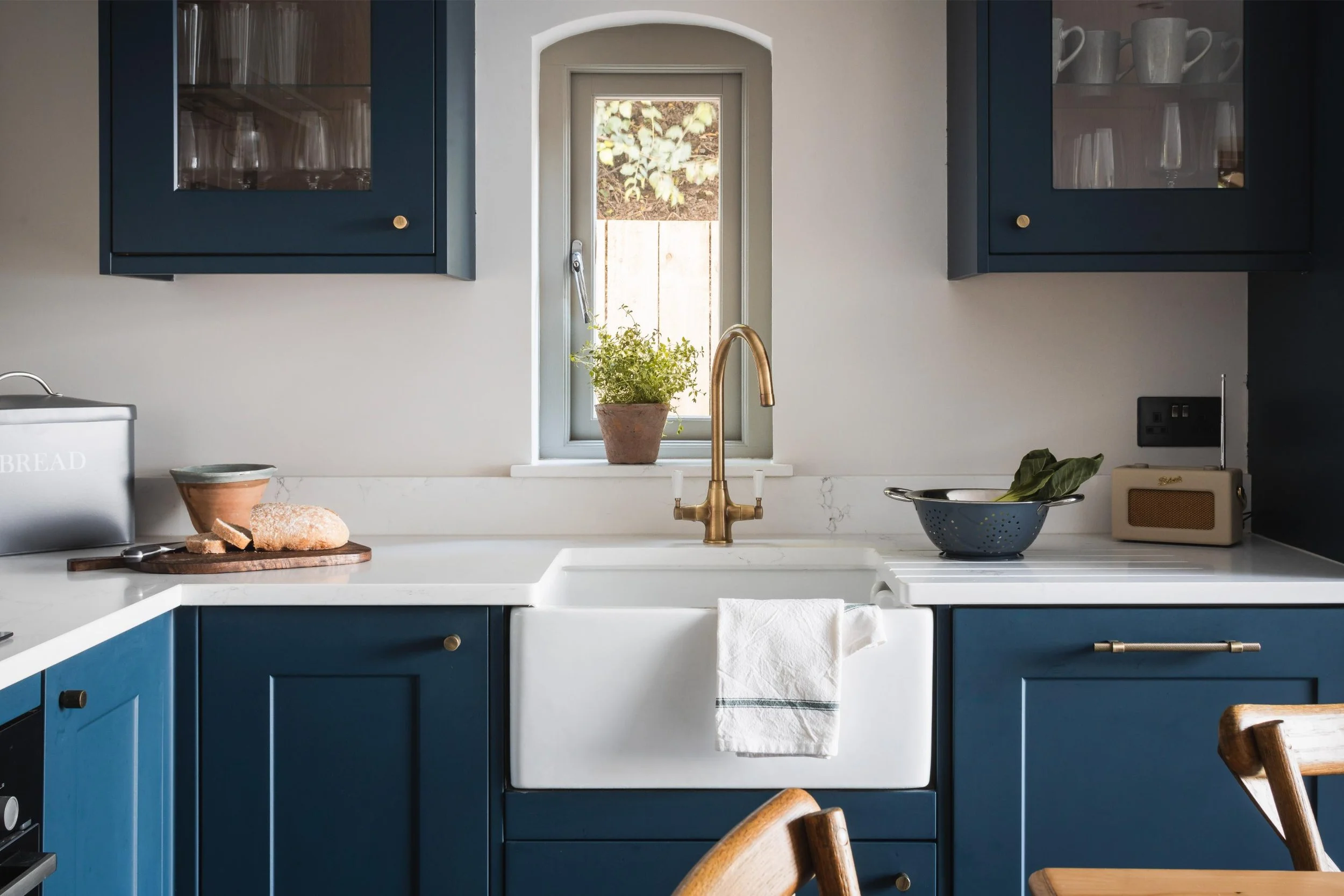 A kitchen with dark blue cabinets, a white marble countertop, and a white farmhouse sink, with a small window above the sink. On the counter, there is a loaf of bread on a wooden cutting board, a small bowl, a potted plant, a colander with leaves, an