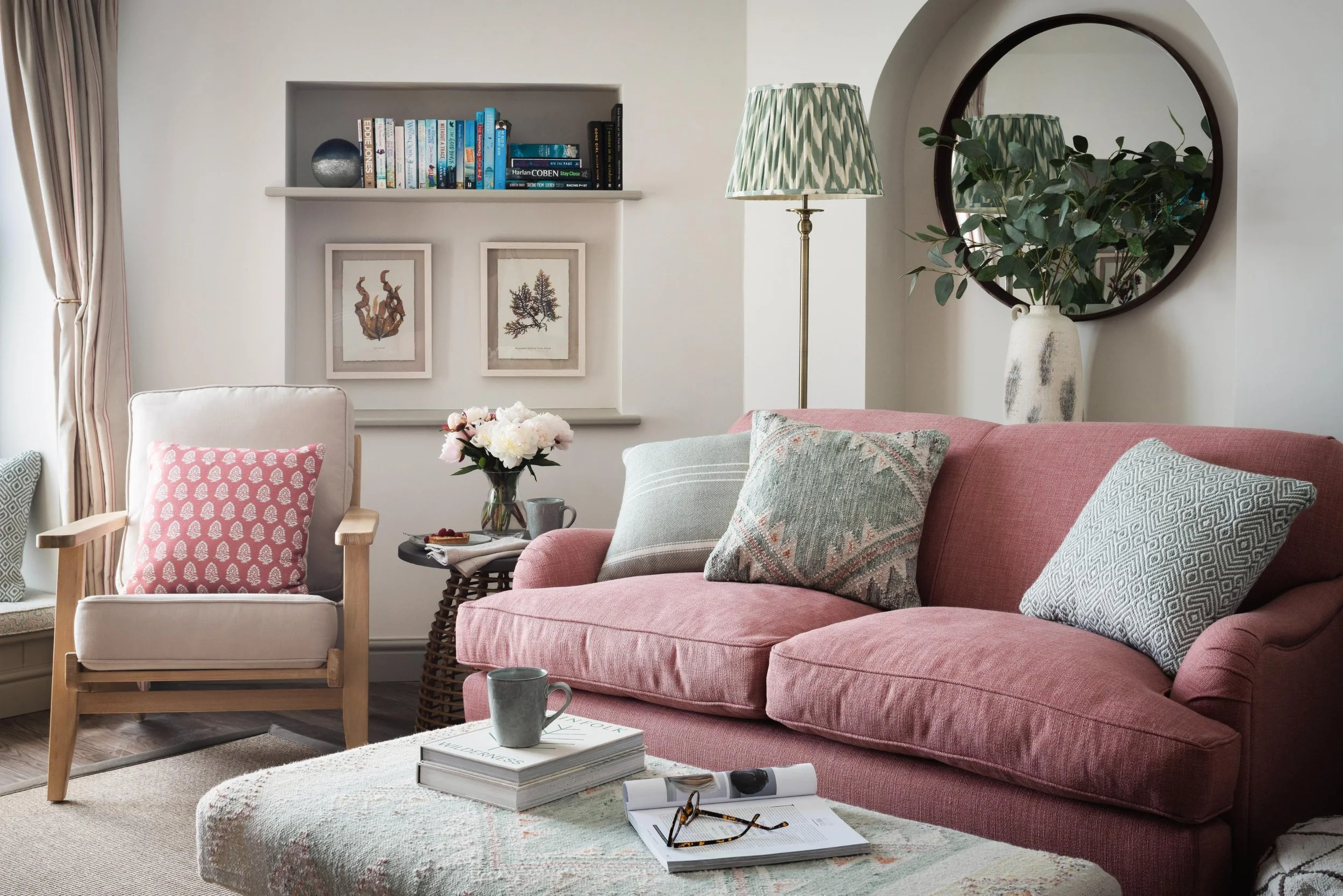 A cozy living room with a pink sofa, an armchair with a pink patterned pillow, a round ottoman with books and magazines, and a side table with a flower vase. There is a bookshelf with books and artwork on the wall, a floor lamp with a patterned shade