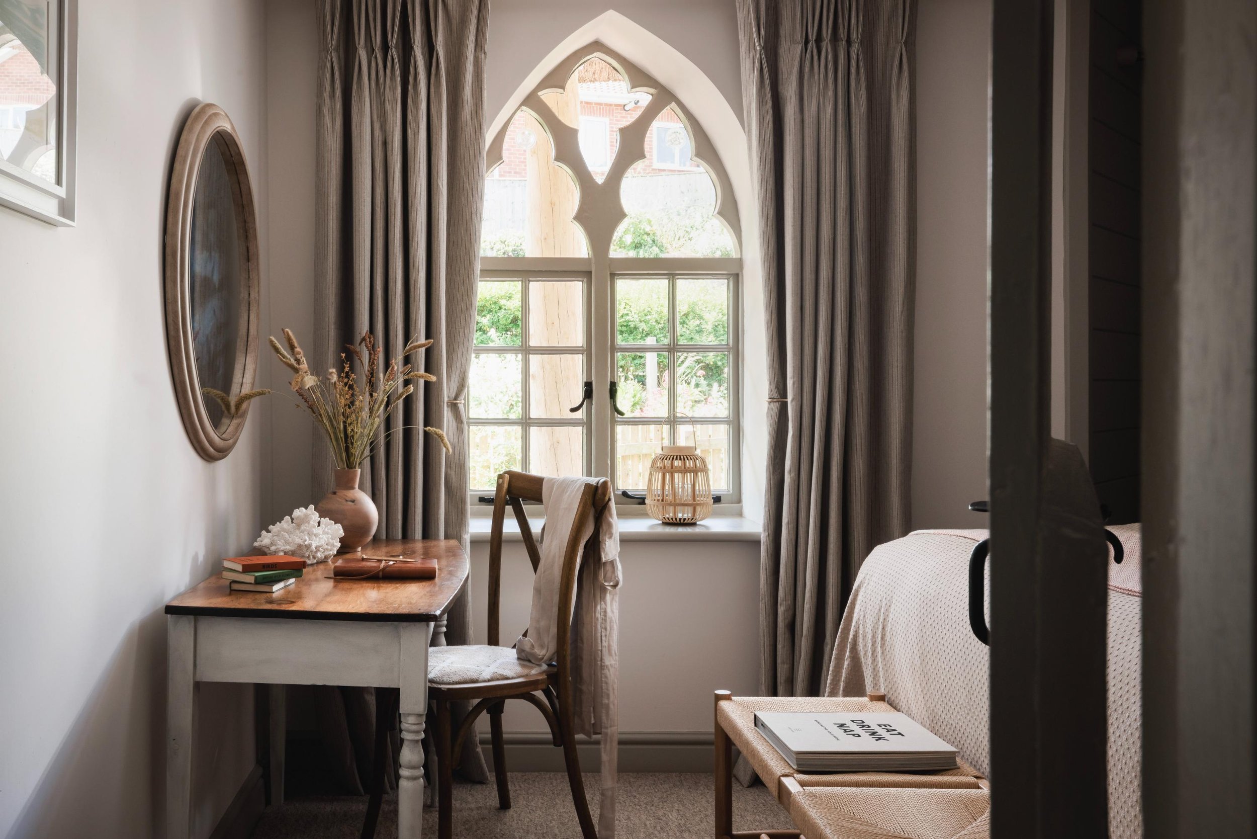 A cozy room with a window that has ivy accents, beige curtains, a small desk with a decorative vase and books, wooden chairs, and a bed with a book on a stool nearby.