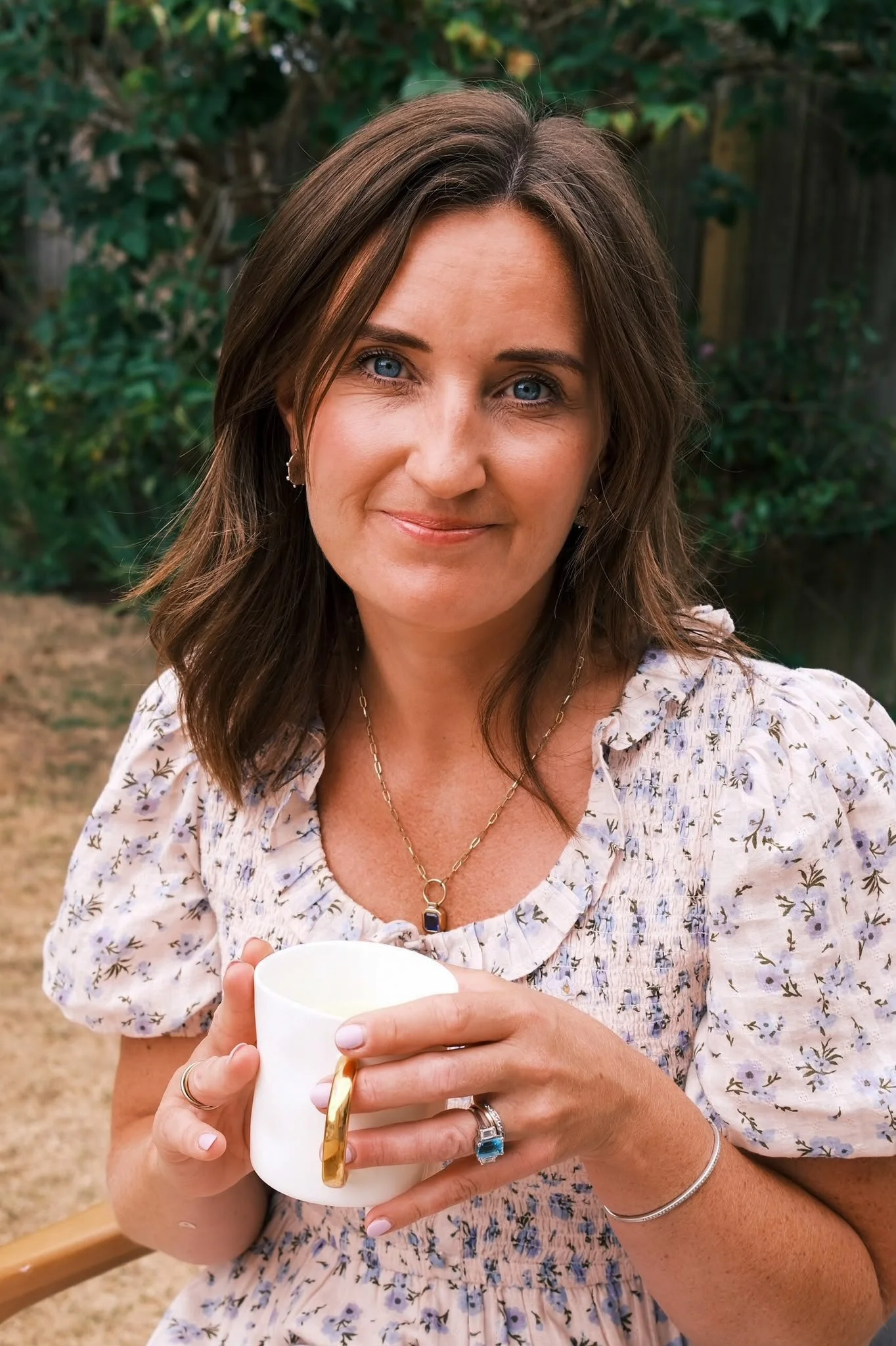 Woman with brown hair and blue eyes holding a white and gold mug outdoors, wearing a floral dress and jewelry.
