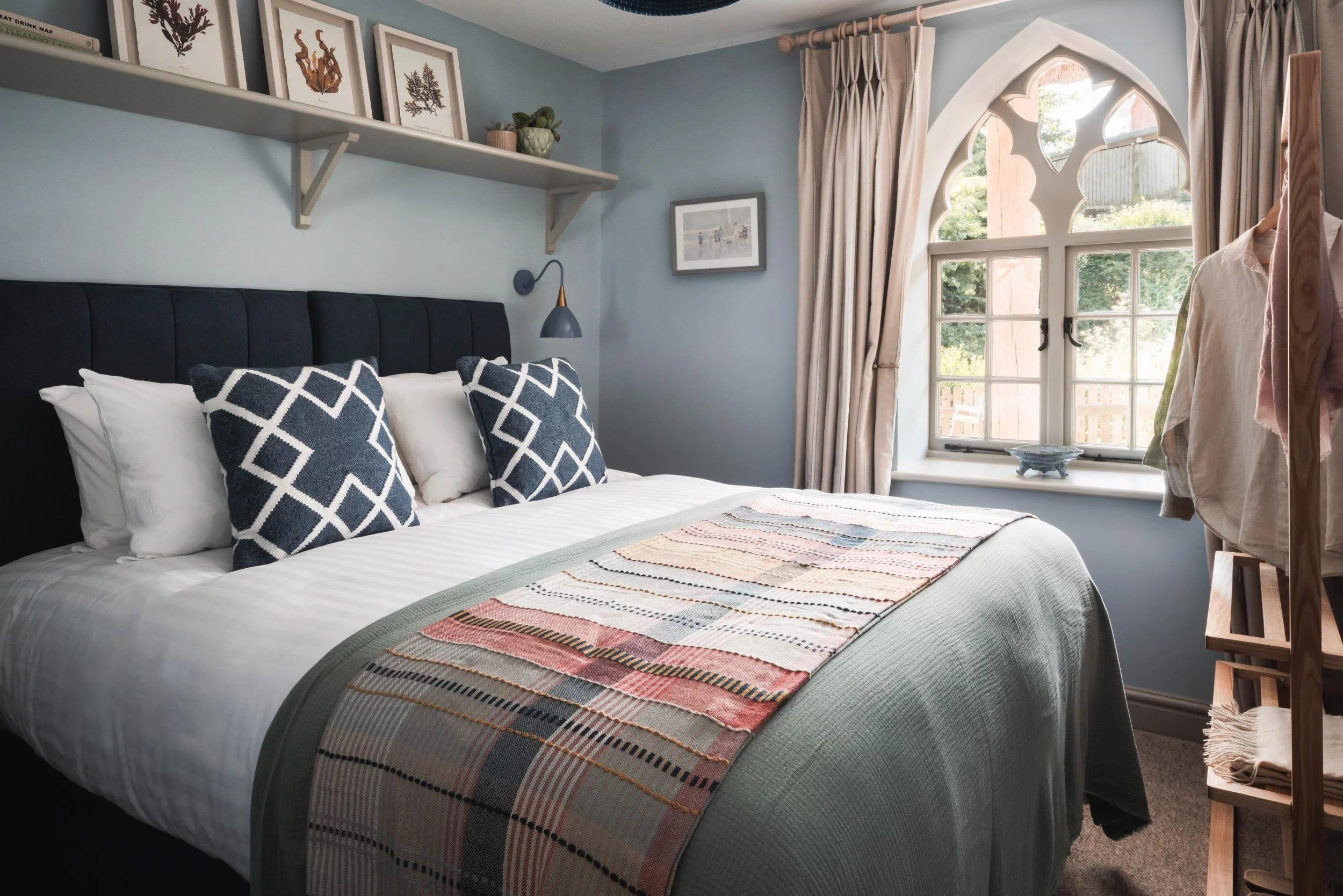 A cozy bedroom with a neatly made bed, dark headboard, white and patterned pillows, a decorative quilted blanket, light blue walls, a floating shelf with framed artwork and potted plants, a large arched window with curtains, and a wooden clothing rac