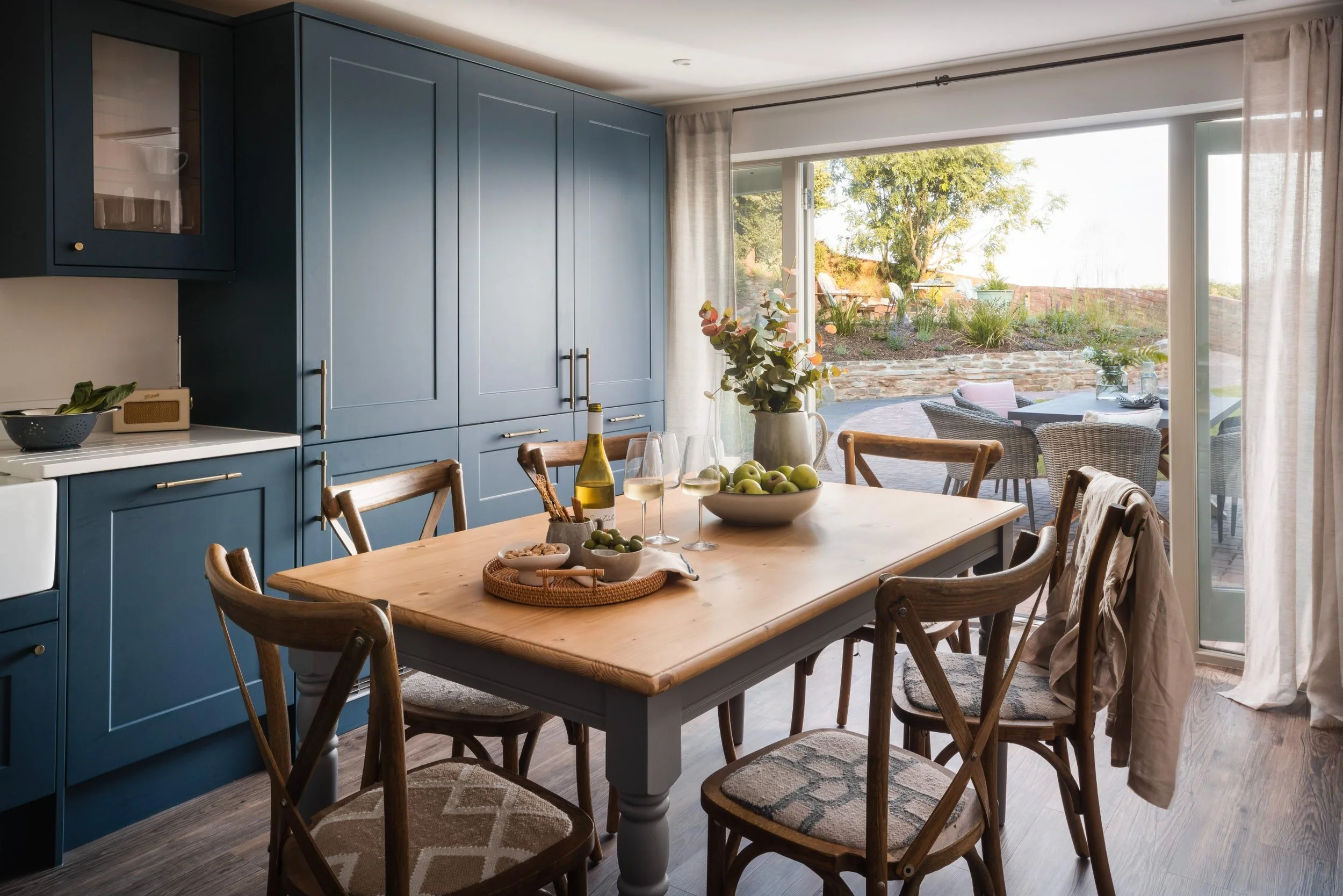 A dining room with a wooden table set with wine glasses, a bowl of green apples, and a plant in a gray vase. Blue cabinets are on the left side, and large sliding glass doors lead outside to a patio with seating and garden greenery.