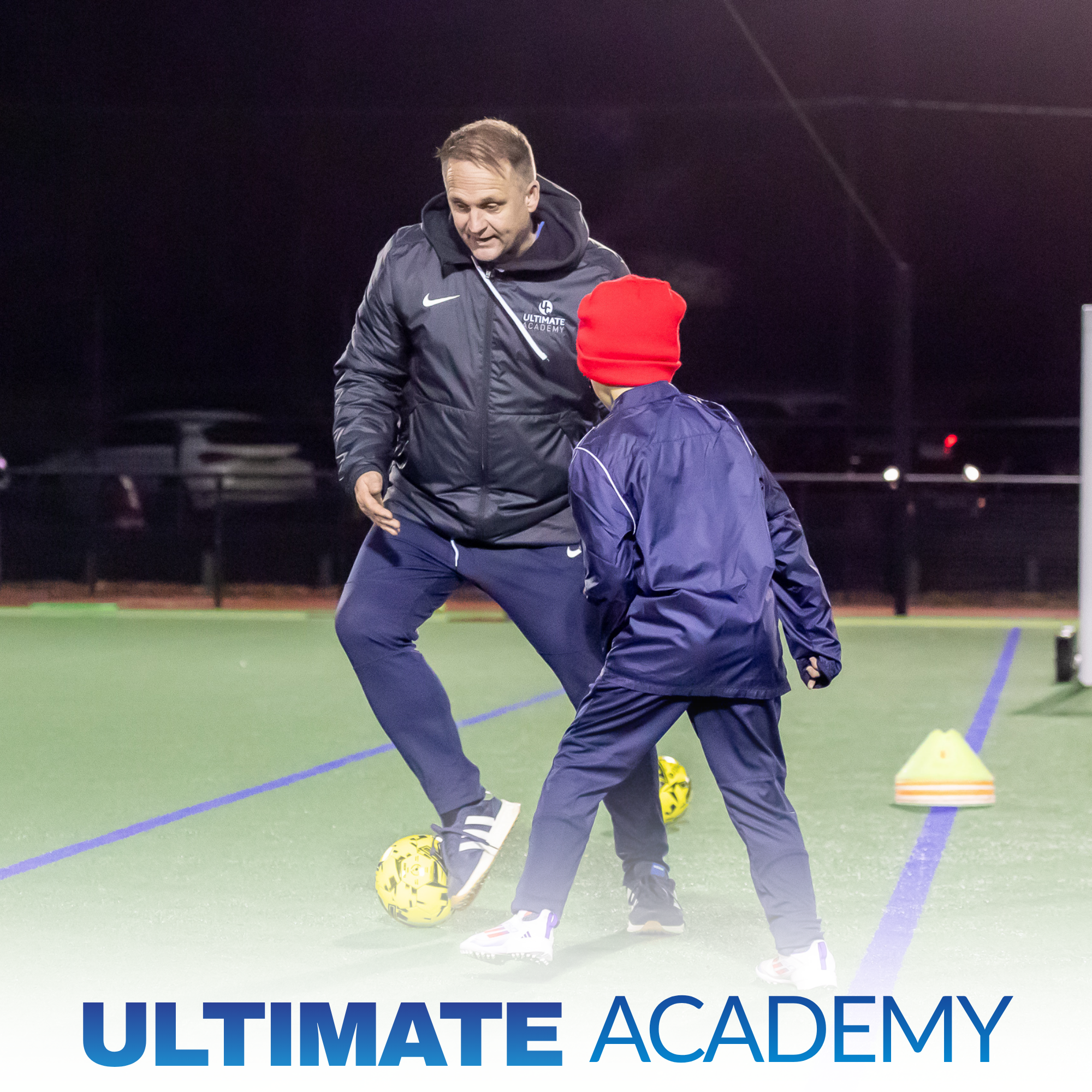 A coach explaining soccer techniques to young players on an indoor field at Ultimate Academy during evening practice.