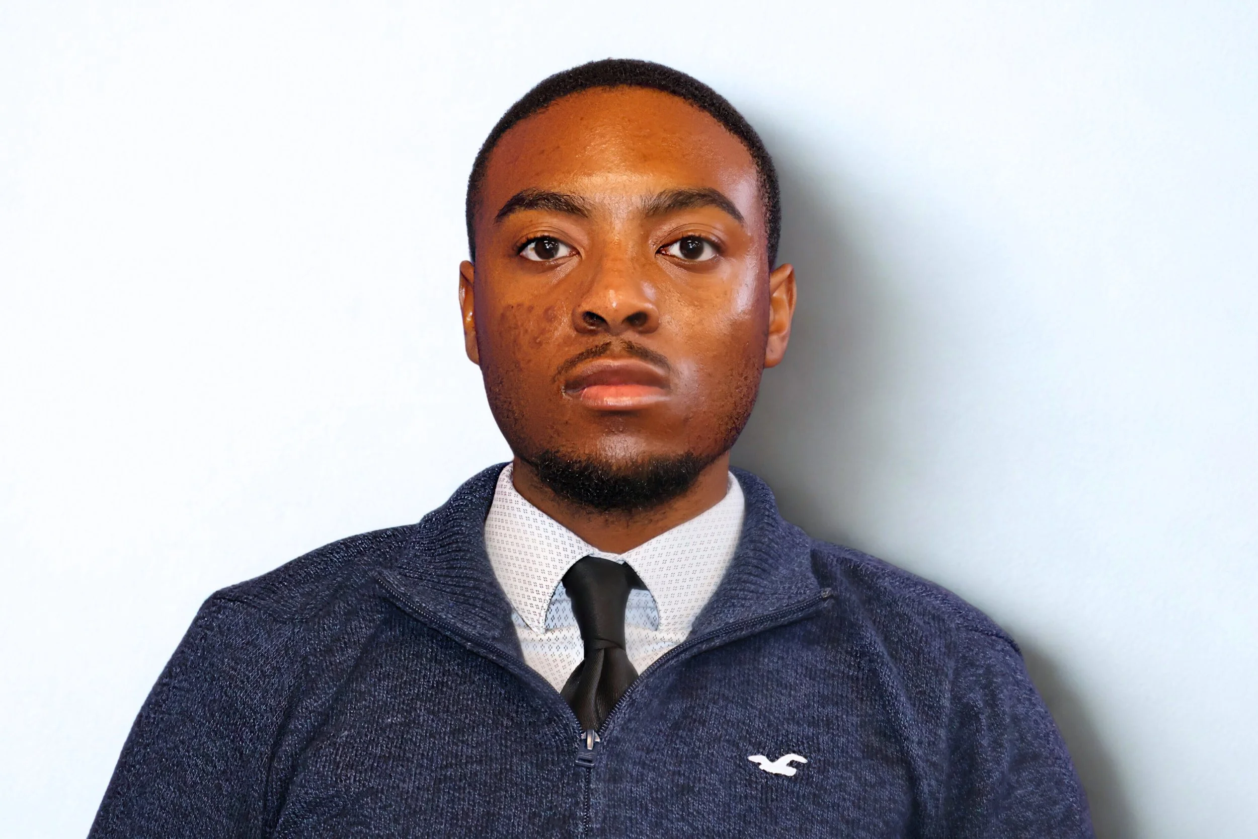 A young man with a serious expression, wearing a blue sweater over a white shirt with a black tie, standing against a plain white background.