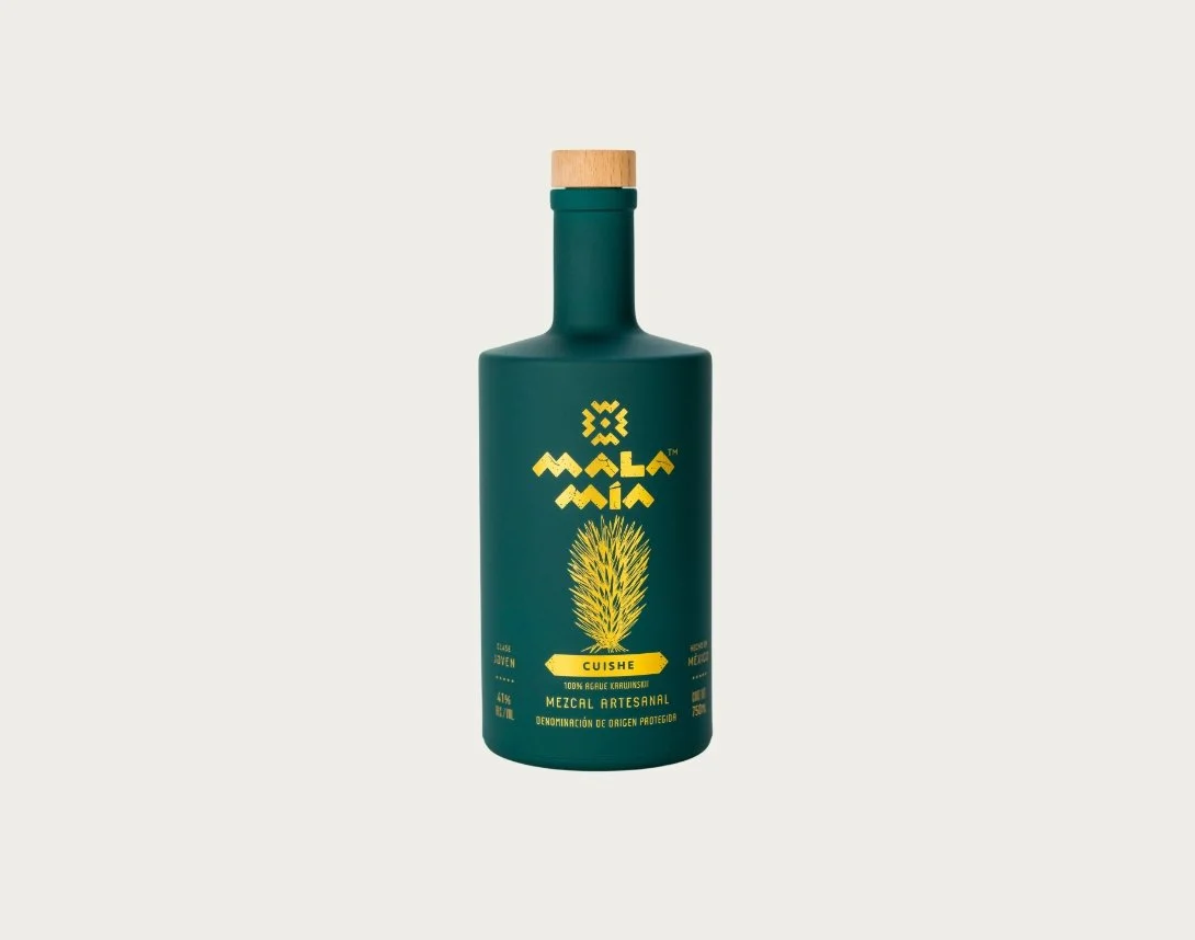 A green bottle of MALA MIA cuishe mezcal with a wooden cap, label featuring a yellow agave plant, and text indicating it is 100% agave, artisanal, and mezcal from Mexico.