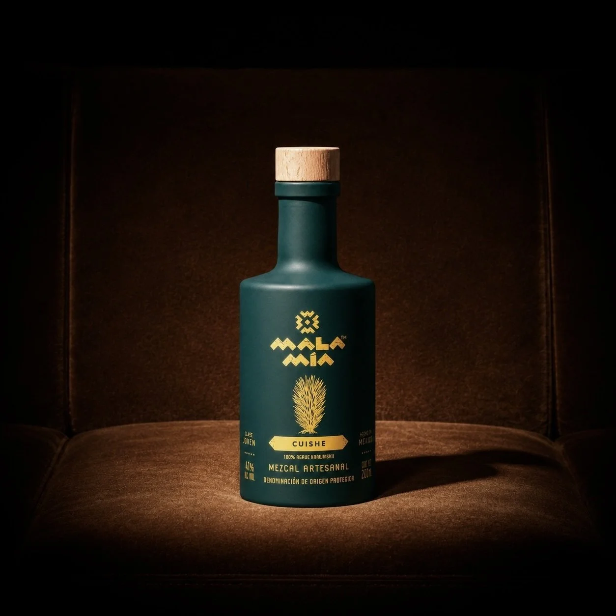 A teal bottle of mezcal artesanal with a wooden cap, featuring a yellow agave plant illustration and text, placed on a cushioned surface with dark background lighting.