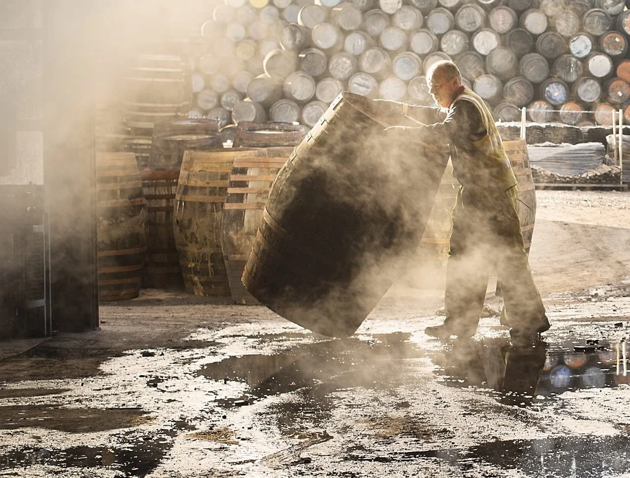 A worker handling a large barrel of fermenting mezcal with steam in the air, surrounded by stacks of barrels.
