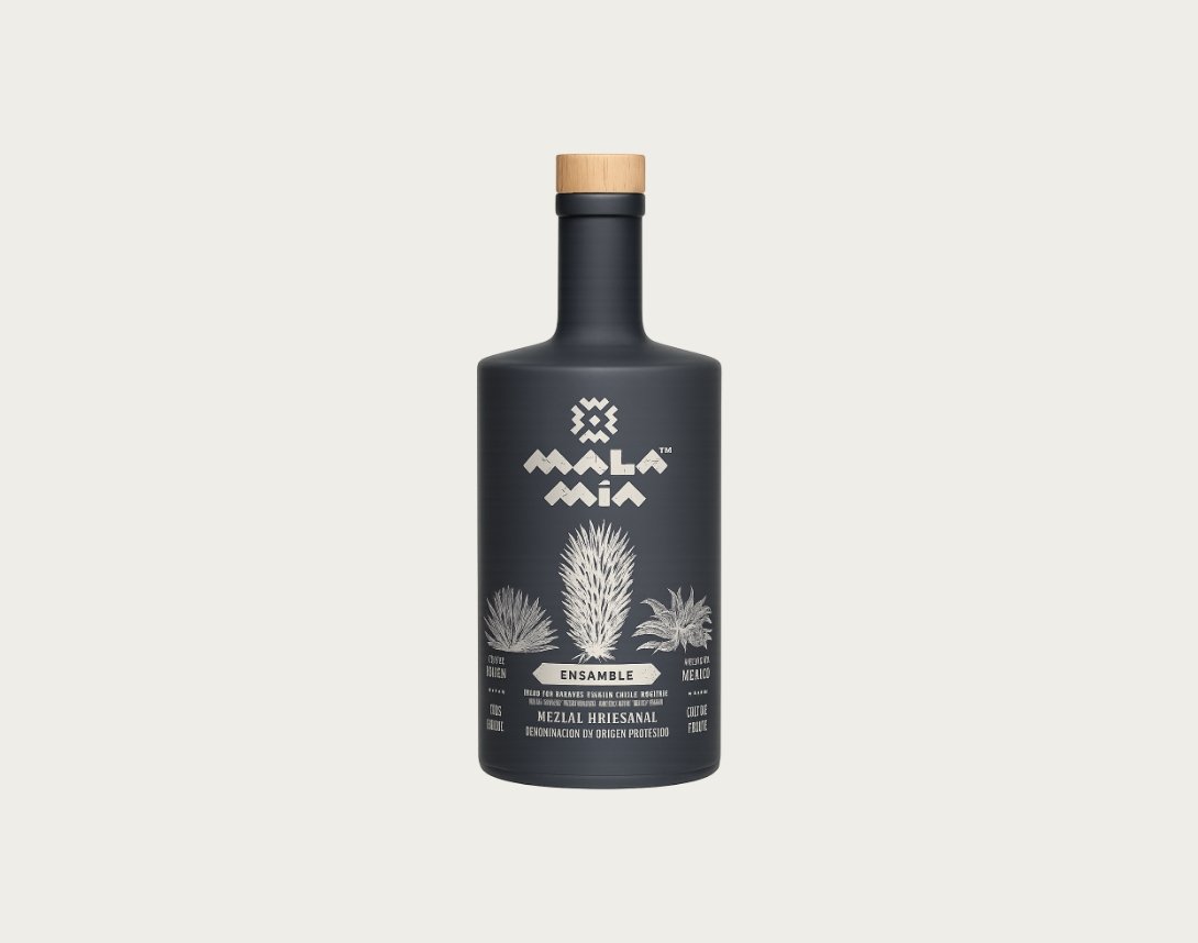 A black bottle of Mala Mía mezcal with a wooden cap, featuring white illustrations of agave plants and text that says this is an ensamble mezcal made with a blend of cuishe, espadín mezcal and tepeztate. 42% alcohol.