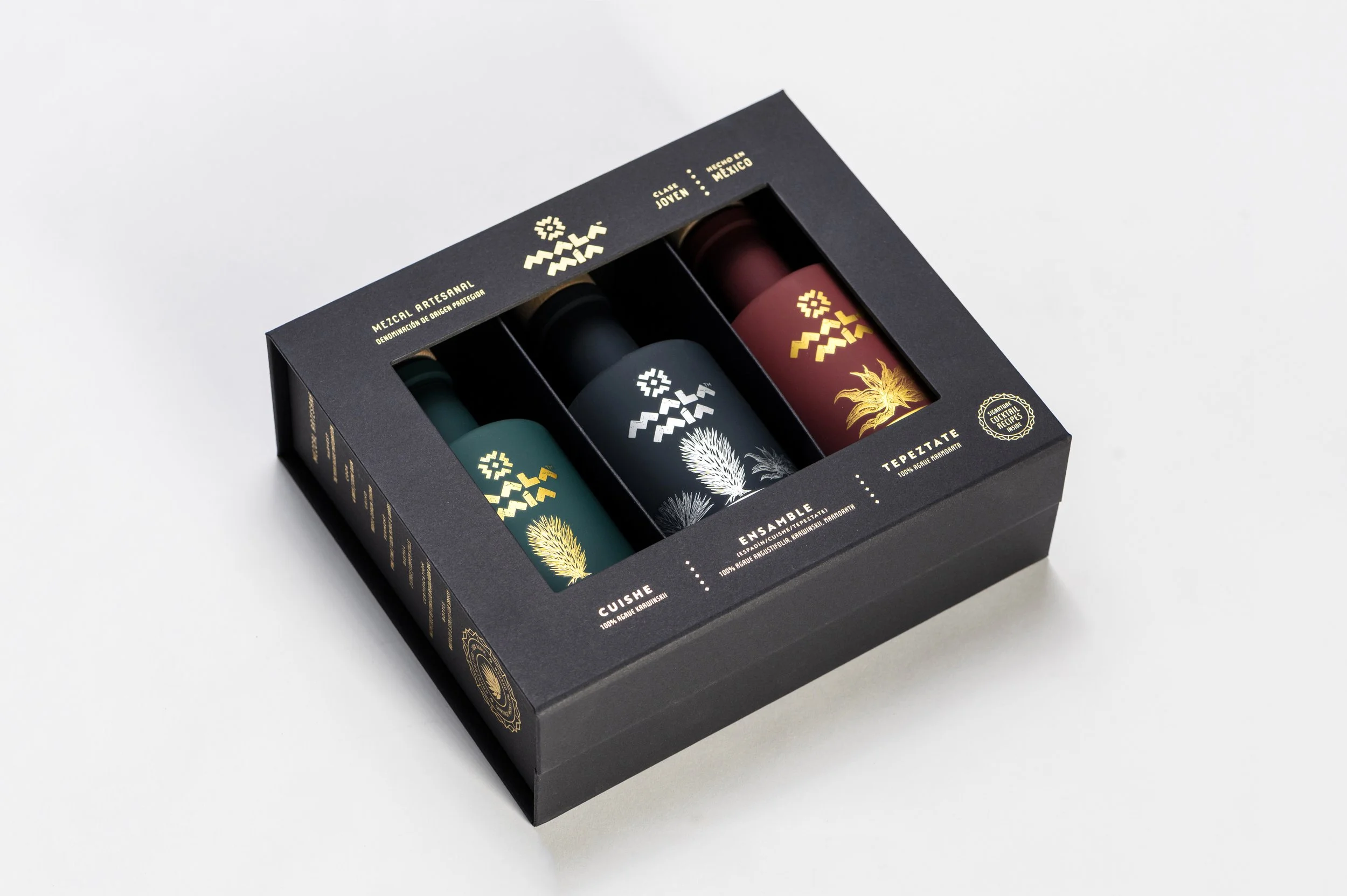 A black box mezcal gift set containing three bottles of artisanal mezcal with colorful designs and gold accents, displayed against a white background.