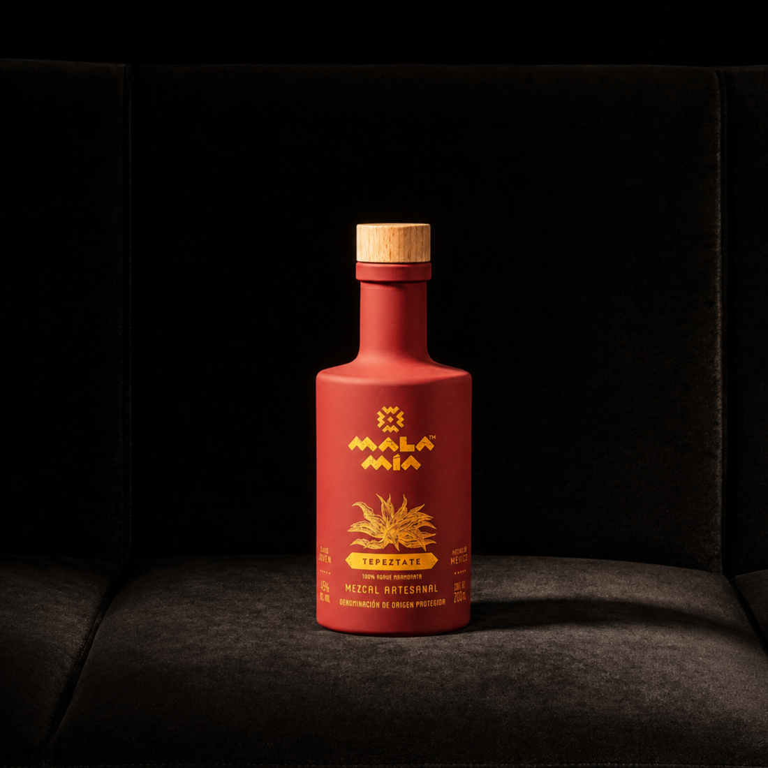 Red bottle of mala mía mezcal tepeztate with yellow label and wooden cap, featuring yellow text and graphics that say this is mezcal from mexico and it is 100% agave, placed on a dark surface against a black background.