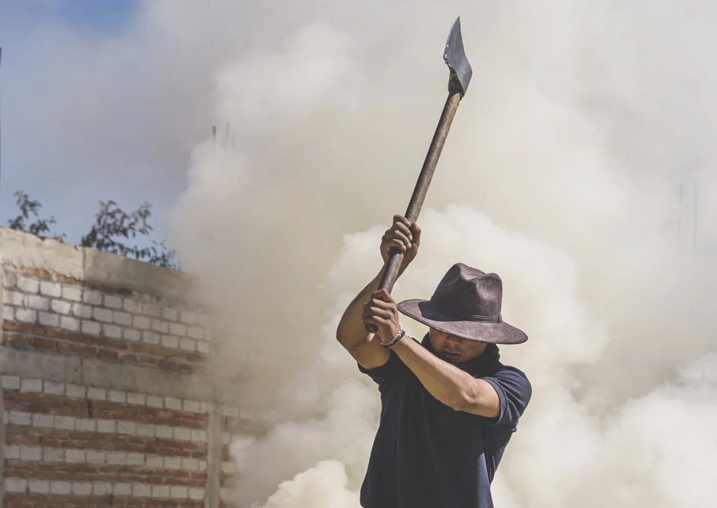 Maestro mezcalero wearing a wide-brimmed hat, holds an axe raised above his head amid smoke, in the process of making mala mia mezcal in oaxaca