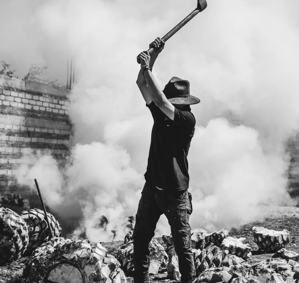 Maestro mezcalero with hat and dark clothing is seen raising an axe above their head, surrounded by smoke and rubble, in the process of making mezcal artesanal.