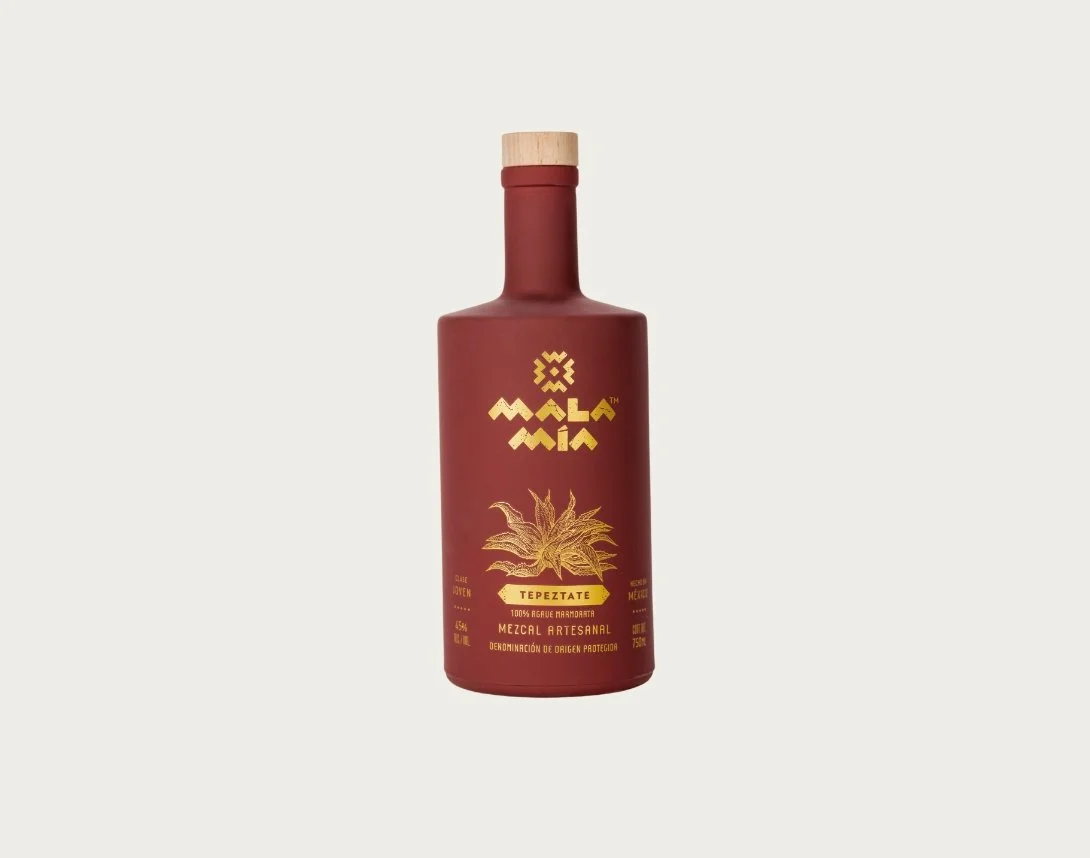 A red bottle of mala mía mezcal with a wooden cap, labeled tepeztate mezcal artesanal containing artisanal mezcal from Mexico with a gold illustration of agave plant.