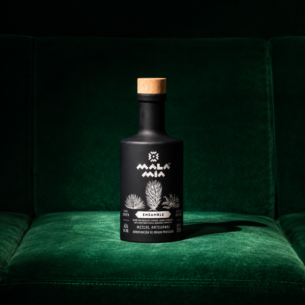 Black bottle of Mala Mia Mezcal artesanal with a wooden cap, sitting on a green velvet seat, featuring a white label with agave plants and text featuring this is mezcal ensamble, made with espadín, cuishe and tepeztate.