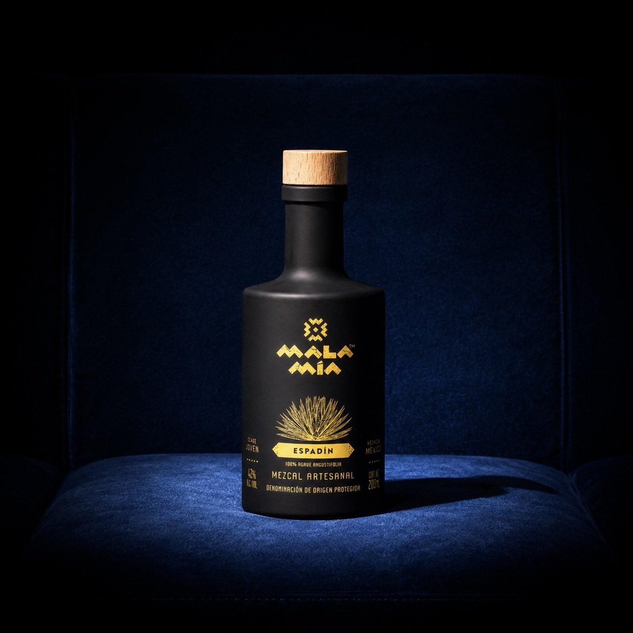 Black bottle of Mala Mía Espadín Mezcal with a wooden cap and yellow labels, including text saying mezcal artesanal, displayed on a blue surface.
