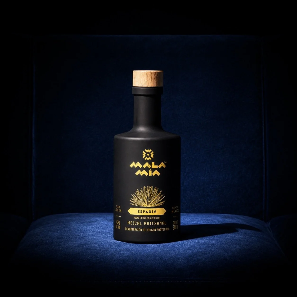 A black bottle of mala mía espadín mezcal with a wooden cap, labeled 'Mezcal Artesanal' placed on a blue cushioned surface, featuring a logo of the 2024 best mezcal of the year san francisco competition winning among the best mezcal brands.