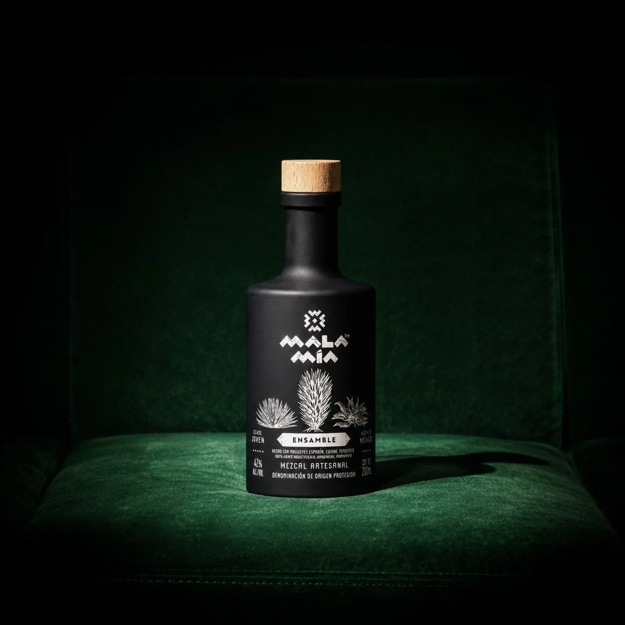 A black bottle of mala mía mezcal with illustrations of three types of agave plants and white text, placed on a green surface.