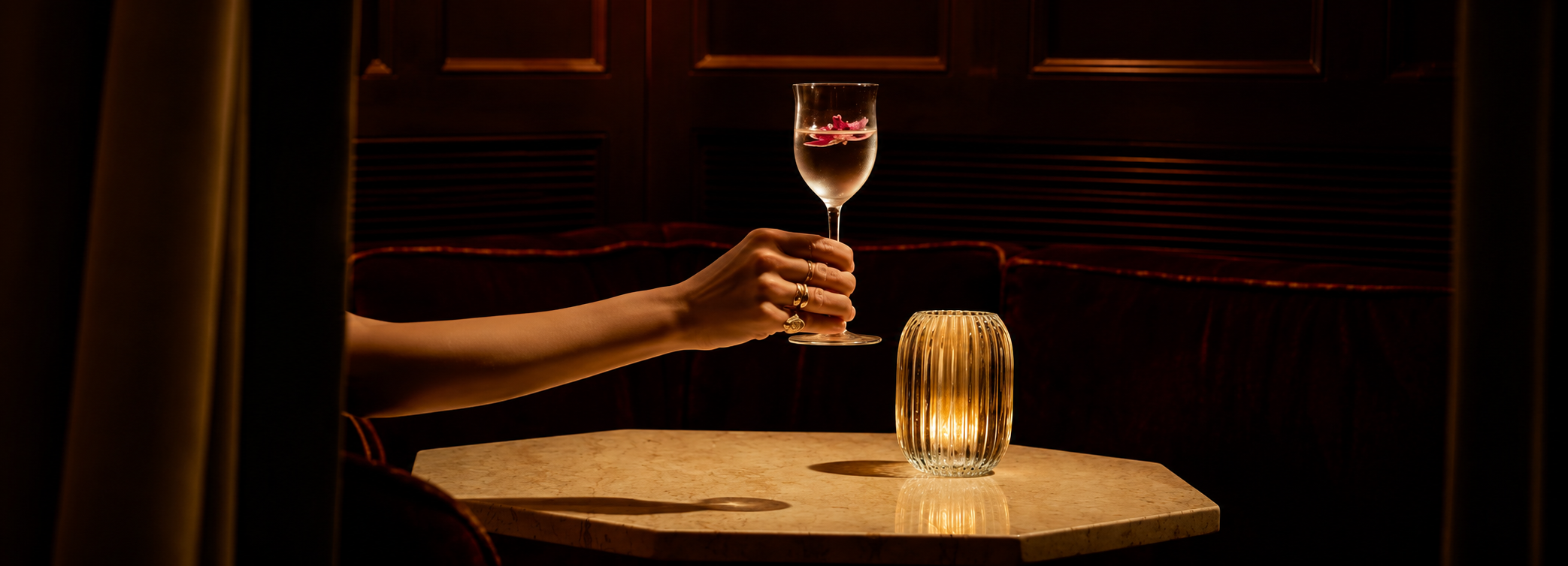 A hand holding a glass of a mezcal cocktail made with espadin mezcal and pink flower petals inside, on a restaurant table with a candle, dark wood-paneled background, and curtains framing the scene.