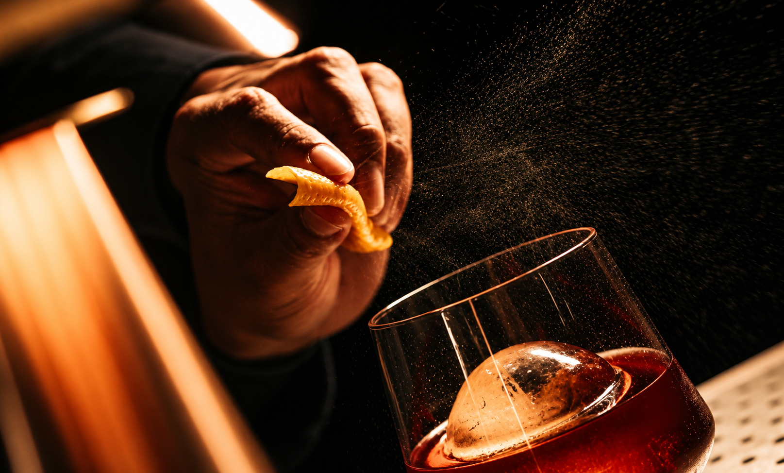 A hand is squeezing lemon into a glass of a mezcal drink with an ice sphere. The scene is dark with warm lighting, and lemon zest is being sprinkled, creating a mist in the air.