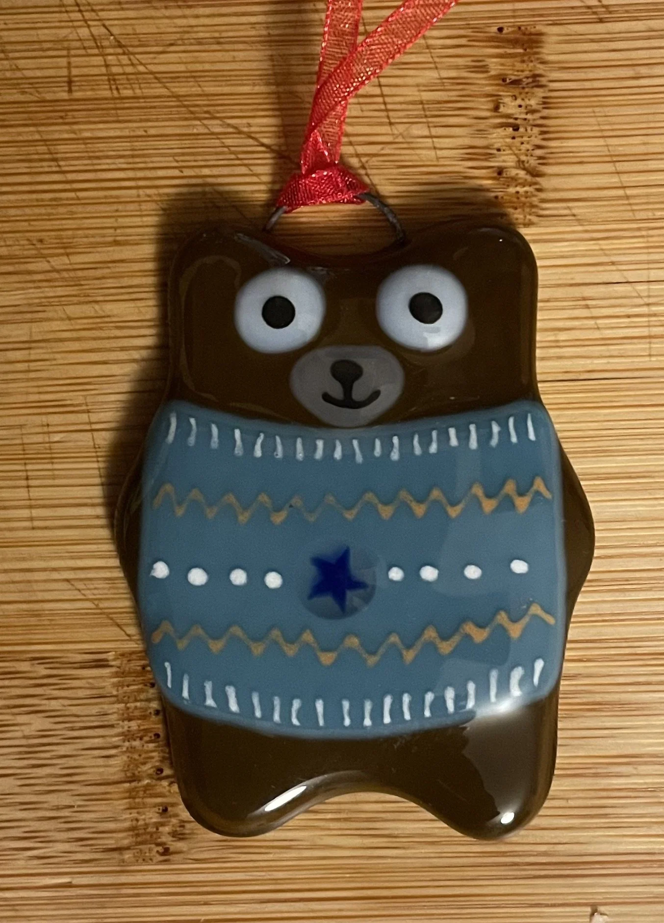 Decorative bear-shaped item with a red ribbon on a wooden surface. The bear has large eyes, a small nose, and a smiling mouth, wearing a blue sweater with star and zigzag patterns.