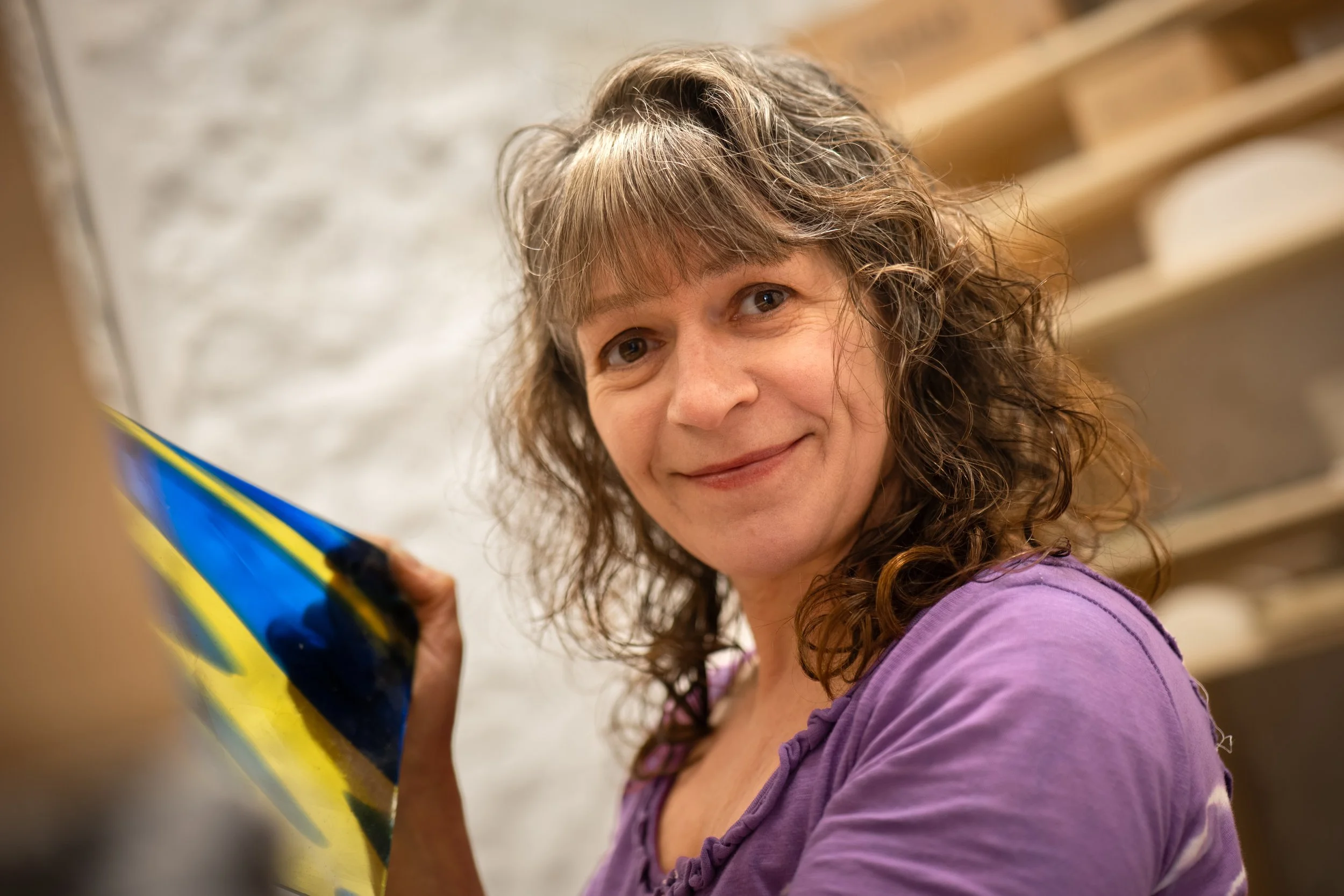 A woman with curly, shoulder-length hair and a purple shirt smiling while holding a blue and yellow skateboard.