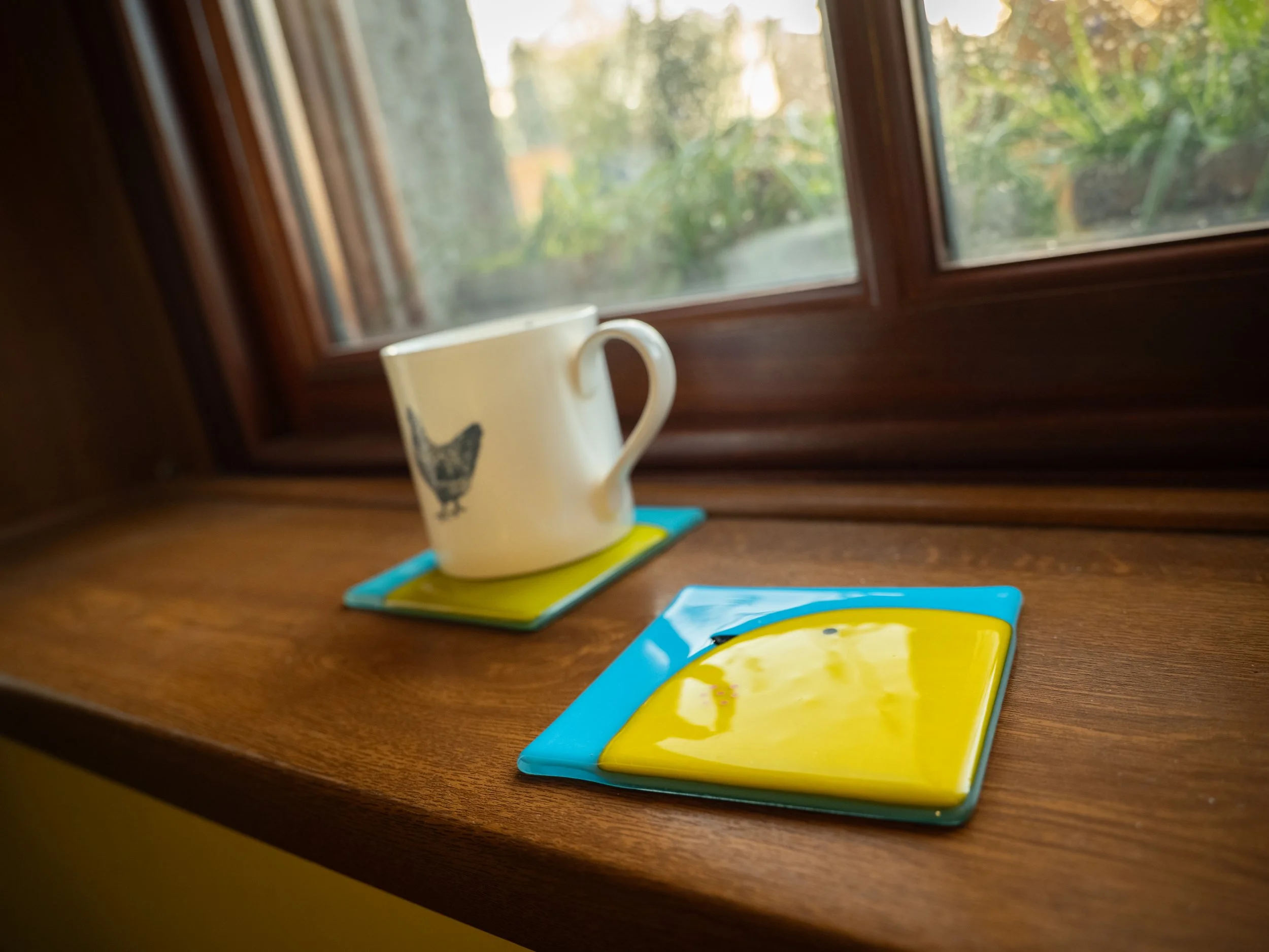 A white coffee mug with a black cat illustration, placed on a colorful tray, on a wooden windowsill with a view of trees outside.