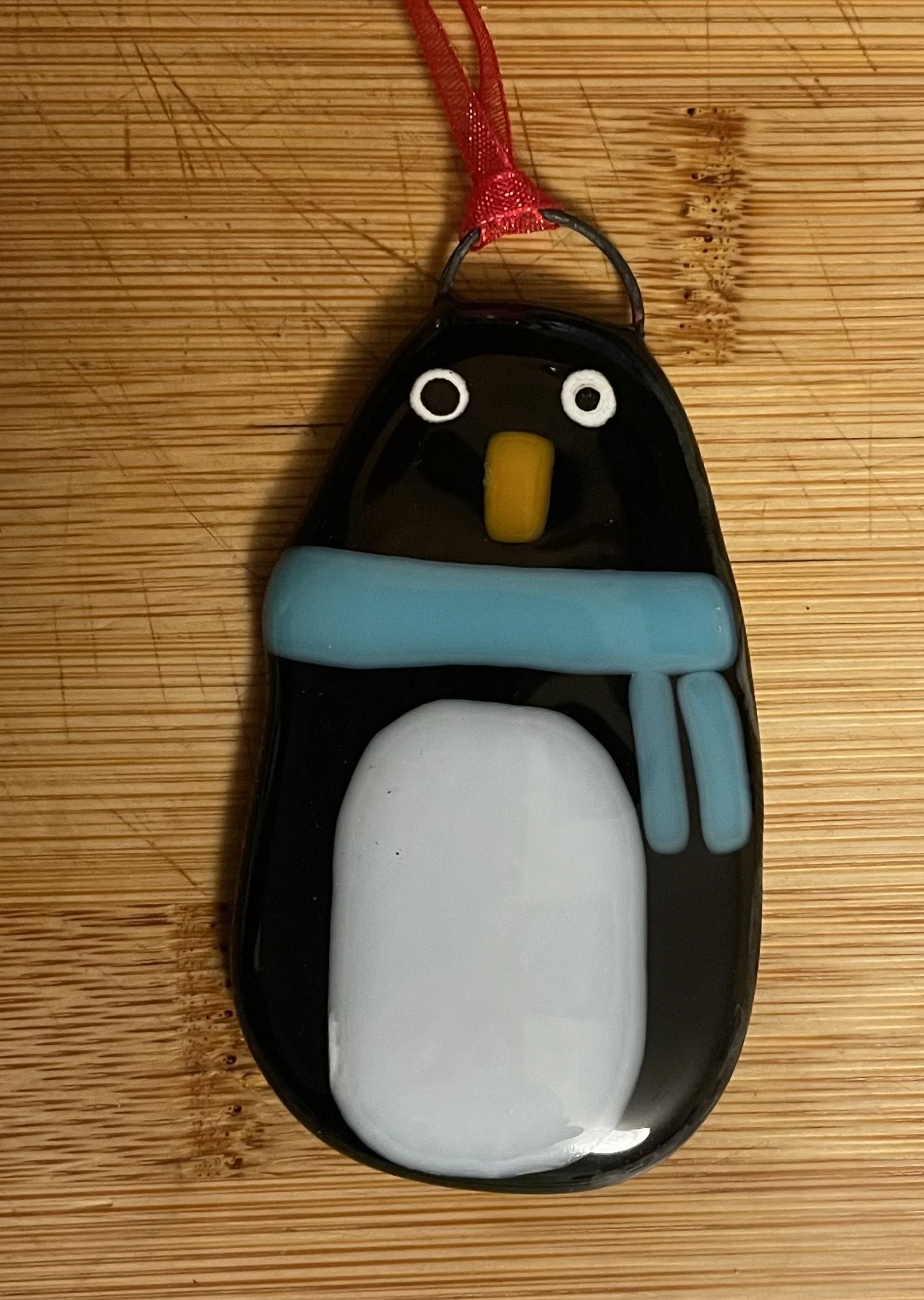 A penguin-shaped air freshener with a red ribbon loop at the top, black body, white belly, yellow beak, and blue wings, placed on a wooden surface.