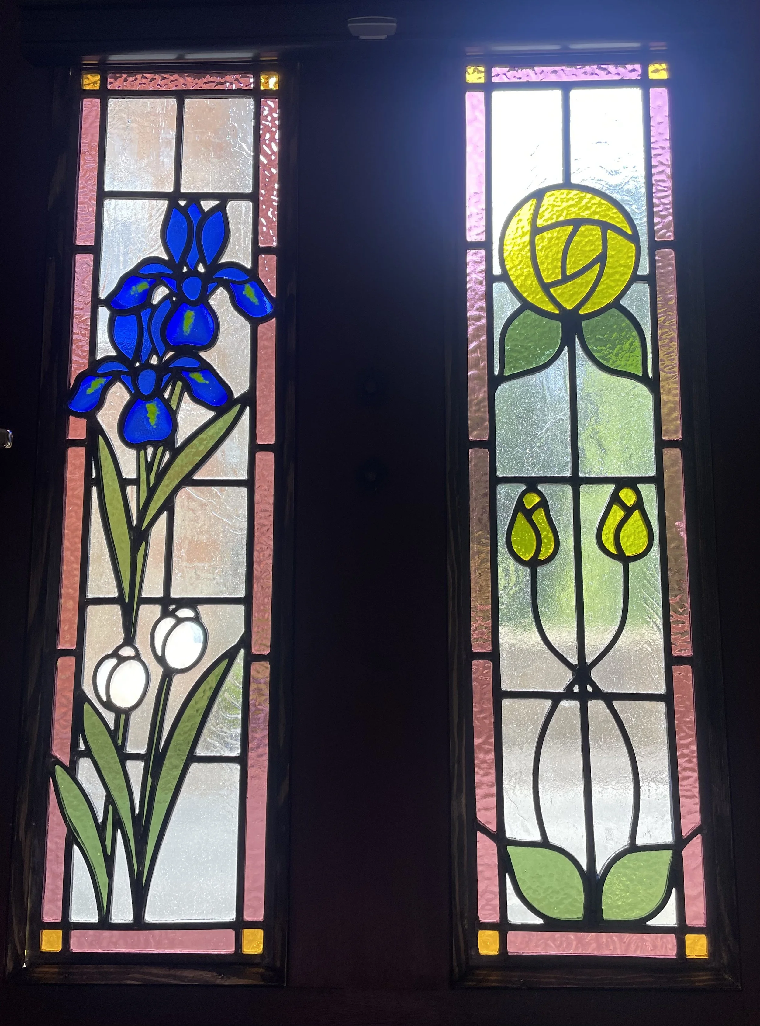Two stained glass windows featuring floral designs. The left window depicts blue irises, and the right window displays a yellow rose with green leaves, separated by dark framing.