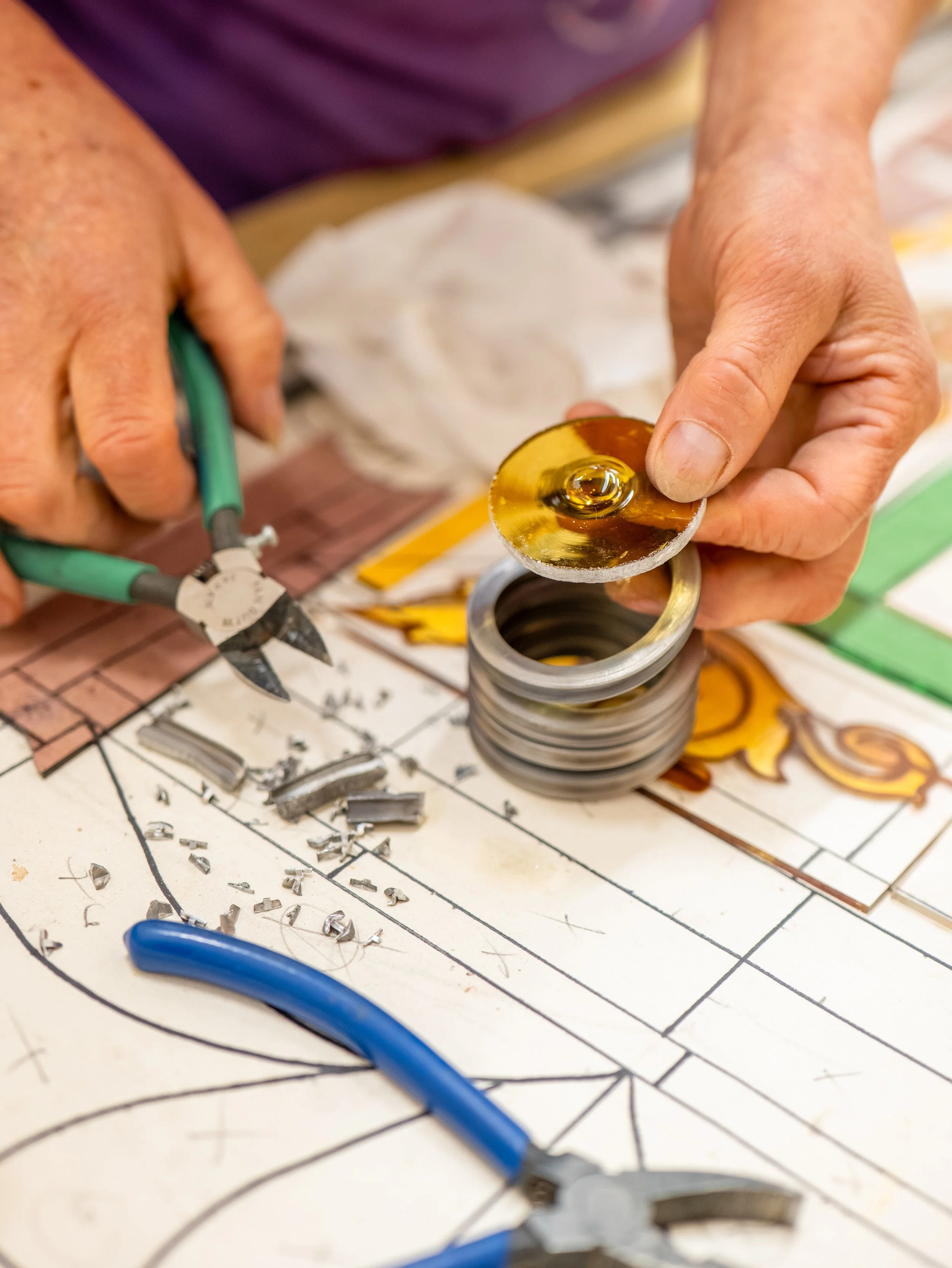 Close-up of someone working on stained glass, holding a small round piece with a yellow and amber design, and tools and glass pieces scattered on a work surface with a pattern.