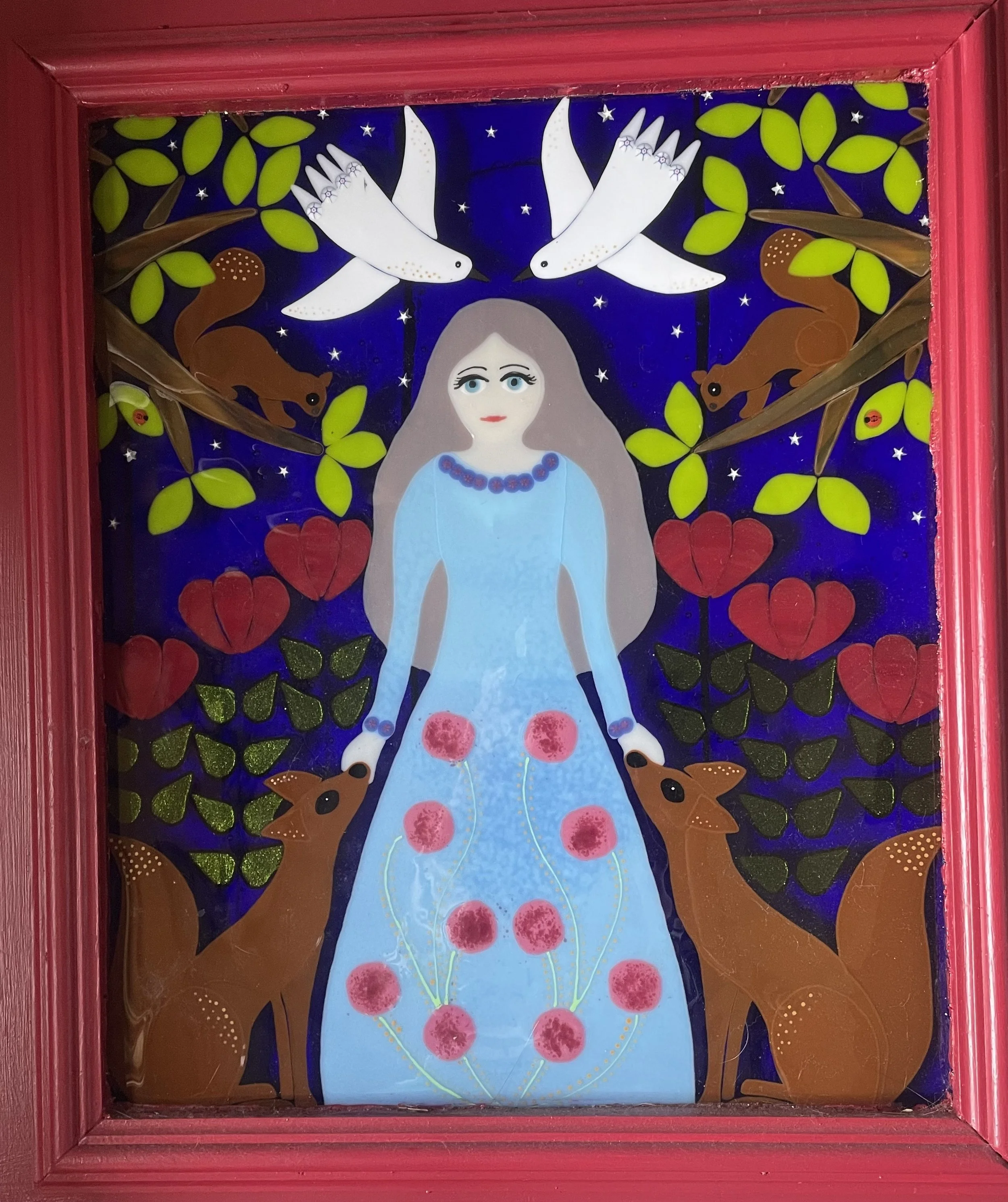 Colorful stained glass artwork depicting a woman with long hair and blue dress, surrounded by white birds, brown animals, and green leaves, with stars and floral patterns in the background, framed with a red wooden border.