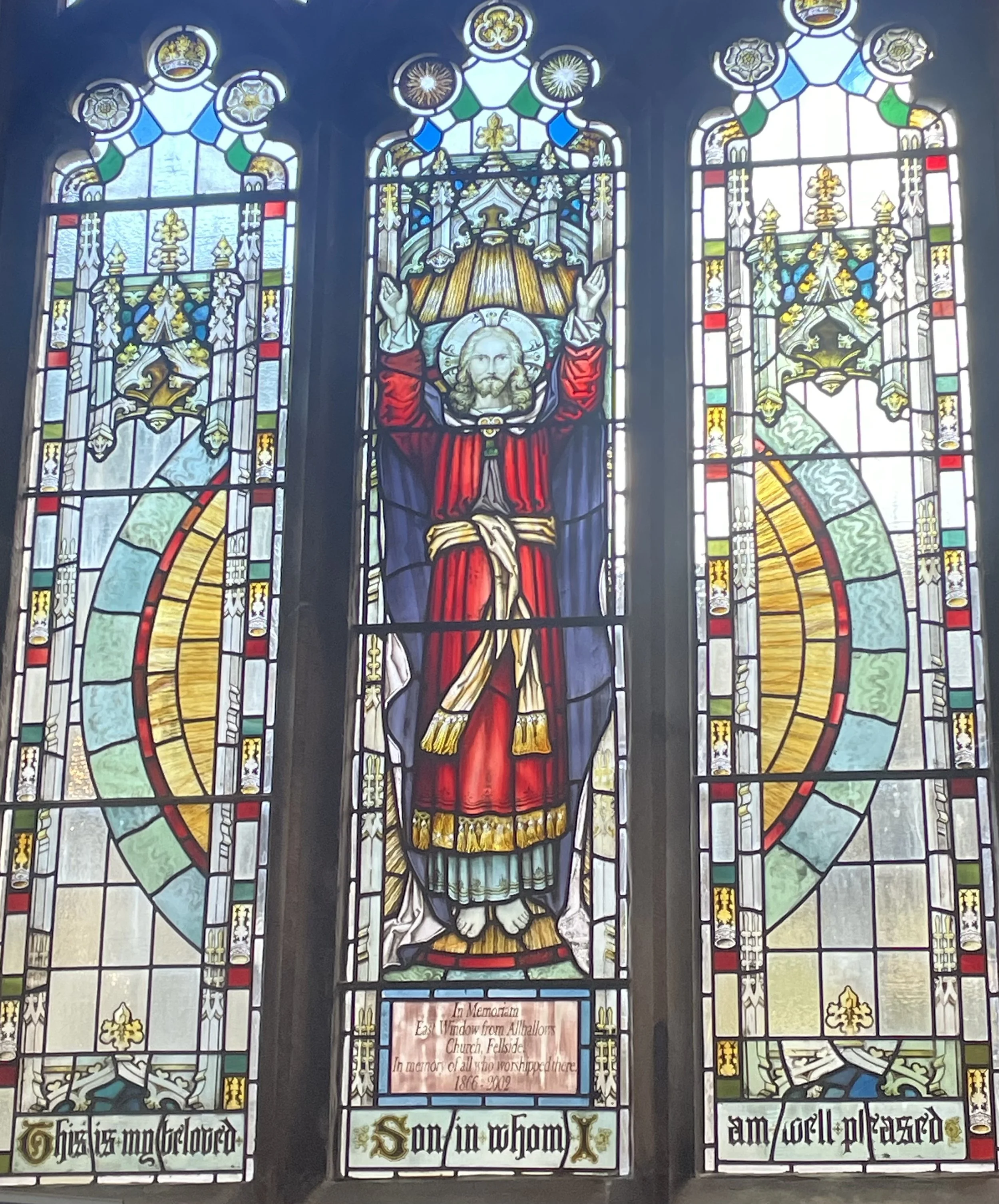 Colorful stained glass window featuring a depiction of Jesus Christ with open hands, a halo, and long hair, surrounded by decorative patterns and symbols, with text at the bottom in blackletter font reading, 'In memory of all who worshipped here 1866