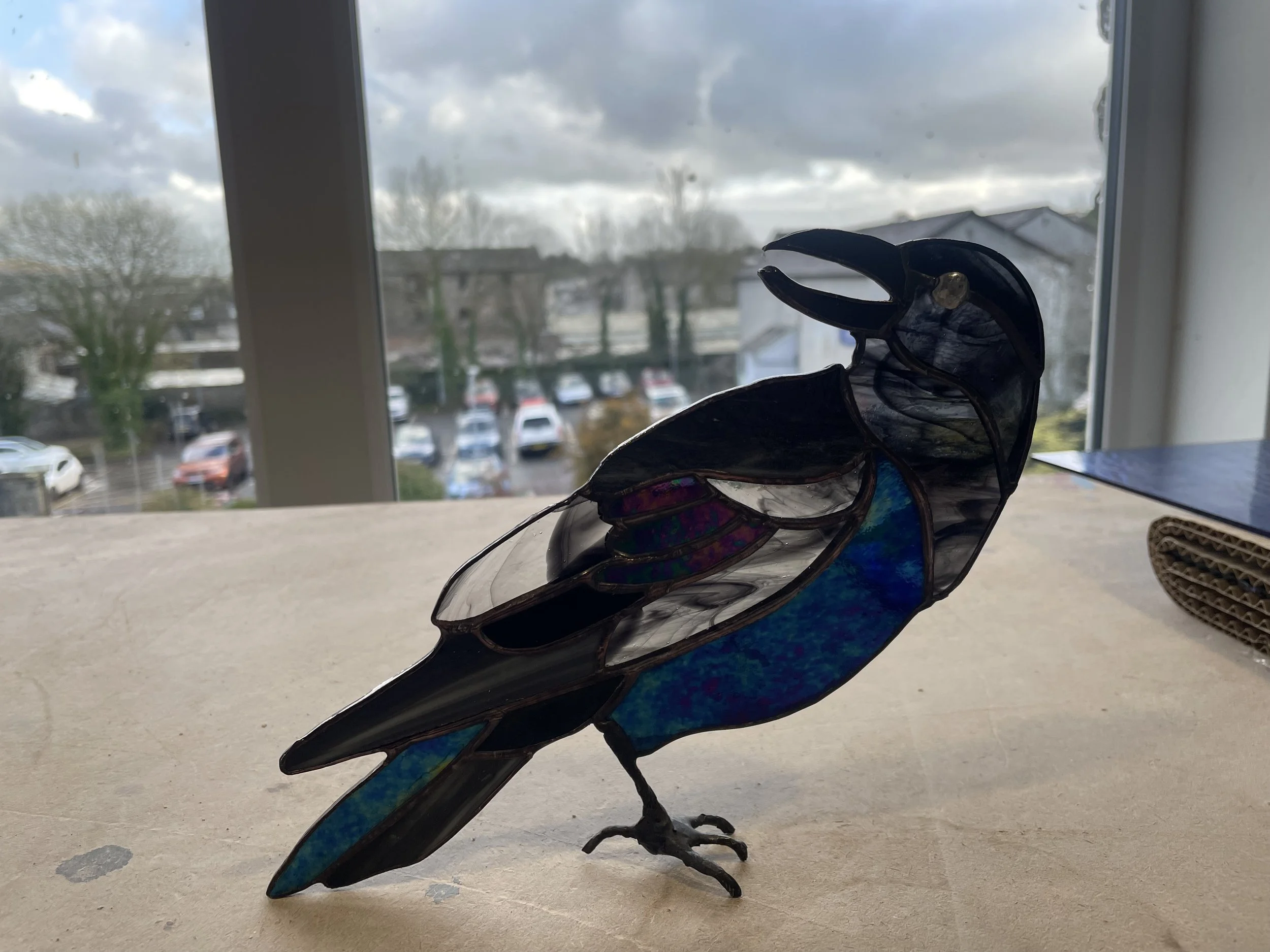 Decorative glass sculpture of a dragon with iridescent colors, positioned on a windowsill with a view of a parking lot and cloudy sky outside.
