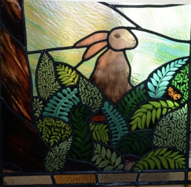 Stained glass window featuring a rabbit among green leaves, with a butterfly and sunlight in the background.