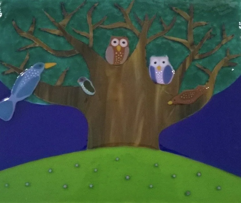 Colorful wall display featuring a tree with hanging owl decorations and painted birds on branches, set against a green hill and purple background.